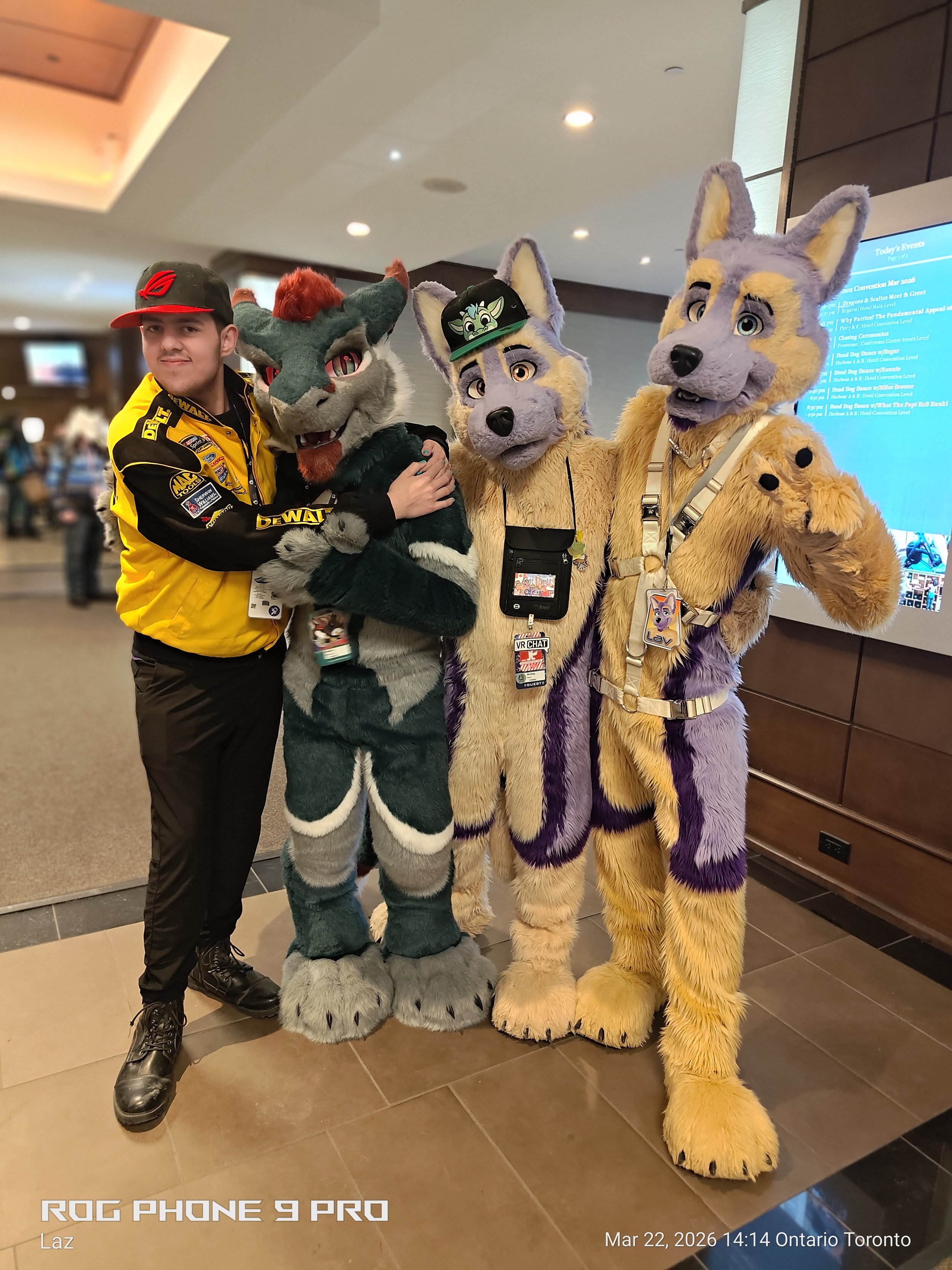 A young man in a black and yellow racing jacket and black cap standing with three people dressed in wolf and fox mascot costumes in an indoor setting with a digital event schedule display in the background.