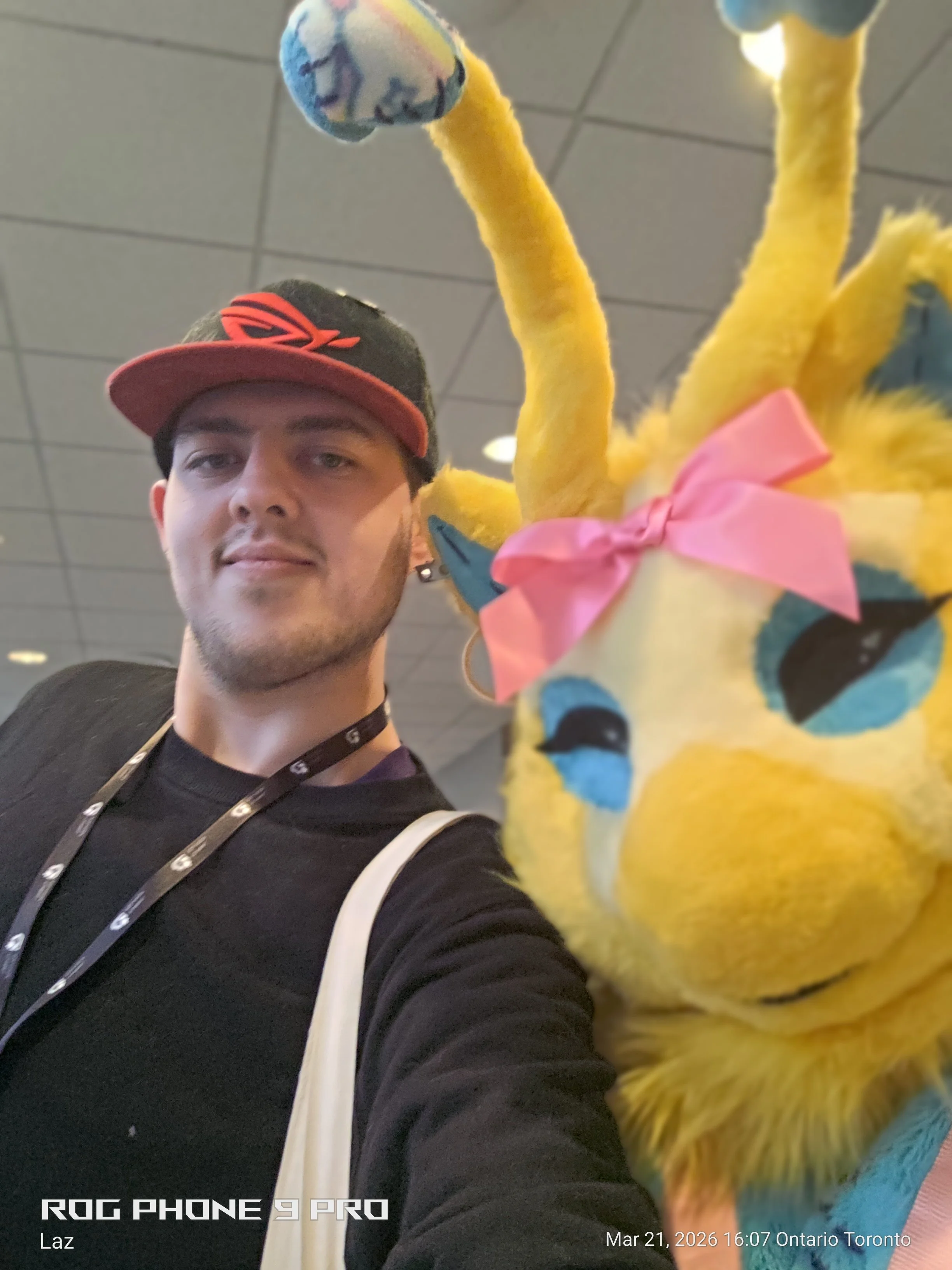 A young man wearing a black baseball cap with K-pop logo, a black shirt, and a lanyard, taking a selfie with a colorful plush toy resembling a bird or a dragon with yellow, blue, and pink accents, large eyes, and pink bow, in an indoor setting with a
