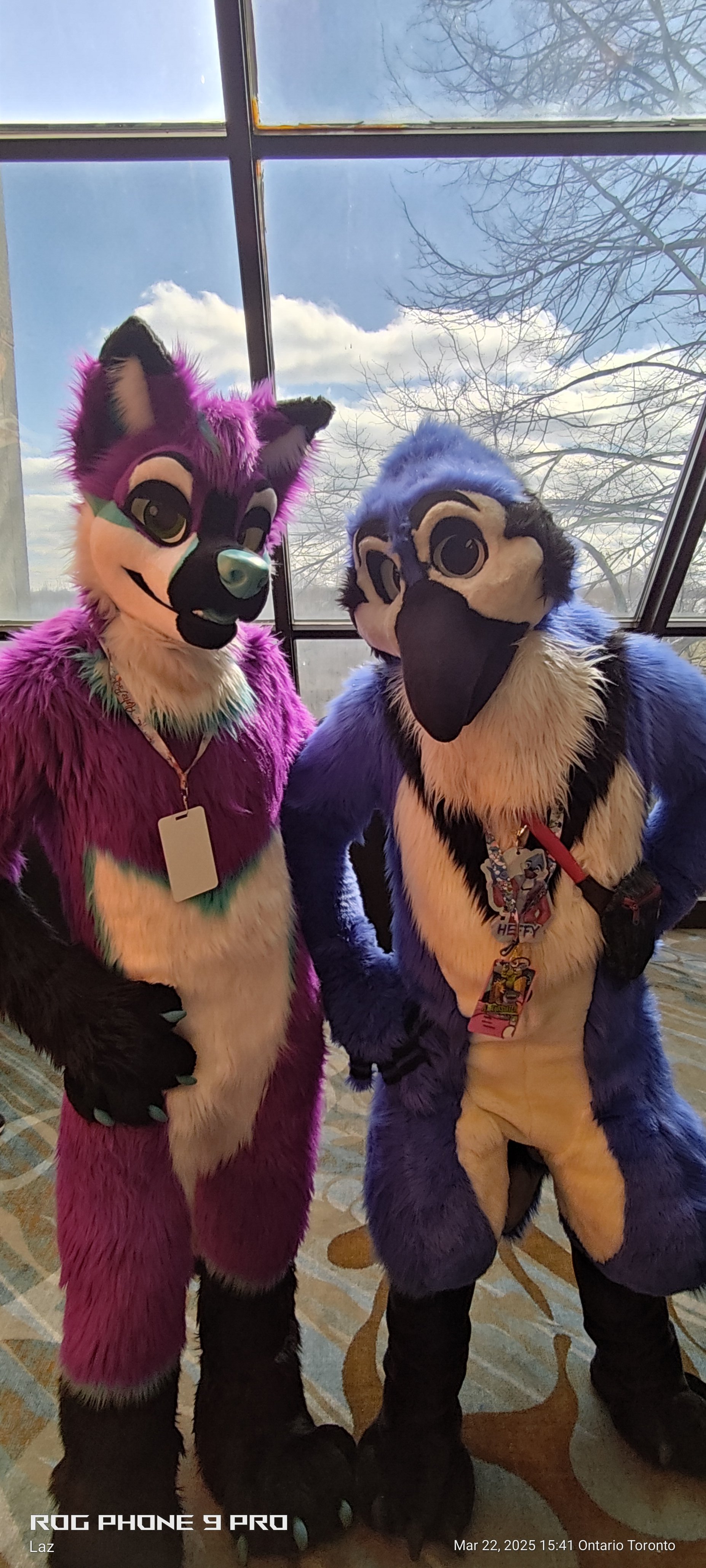 Two people dressed in colorful furry animal costumes, a purple fox and a blue and white bird, standing indoors near a large window with a view of trees and blue sky.