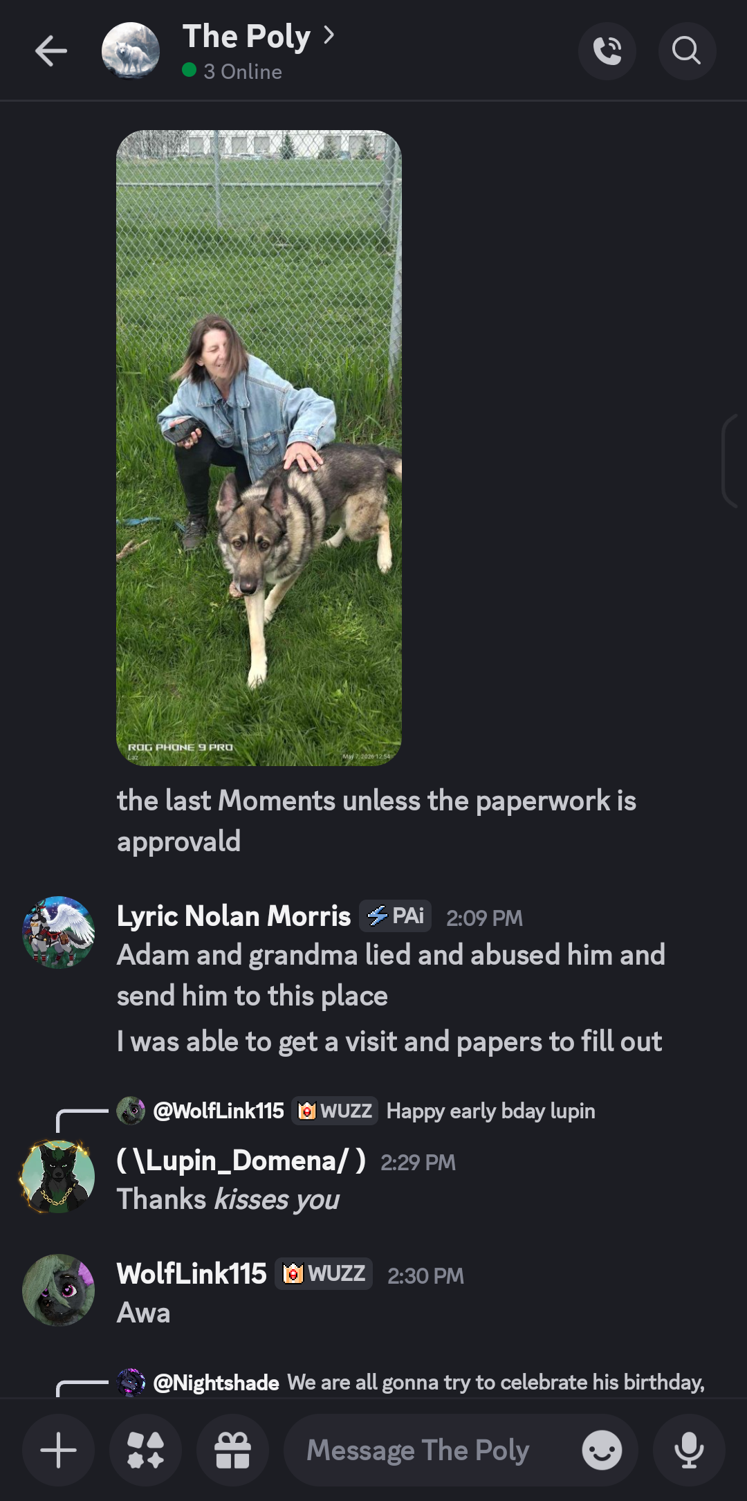 A woman in a denim jacket crouching next to a large German Shepherd dog on a grassy field near a chain-link fence.