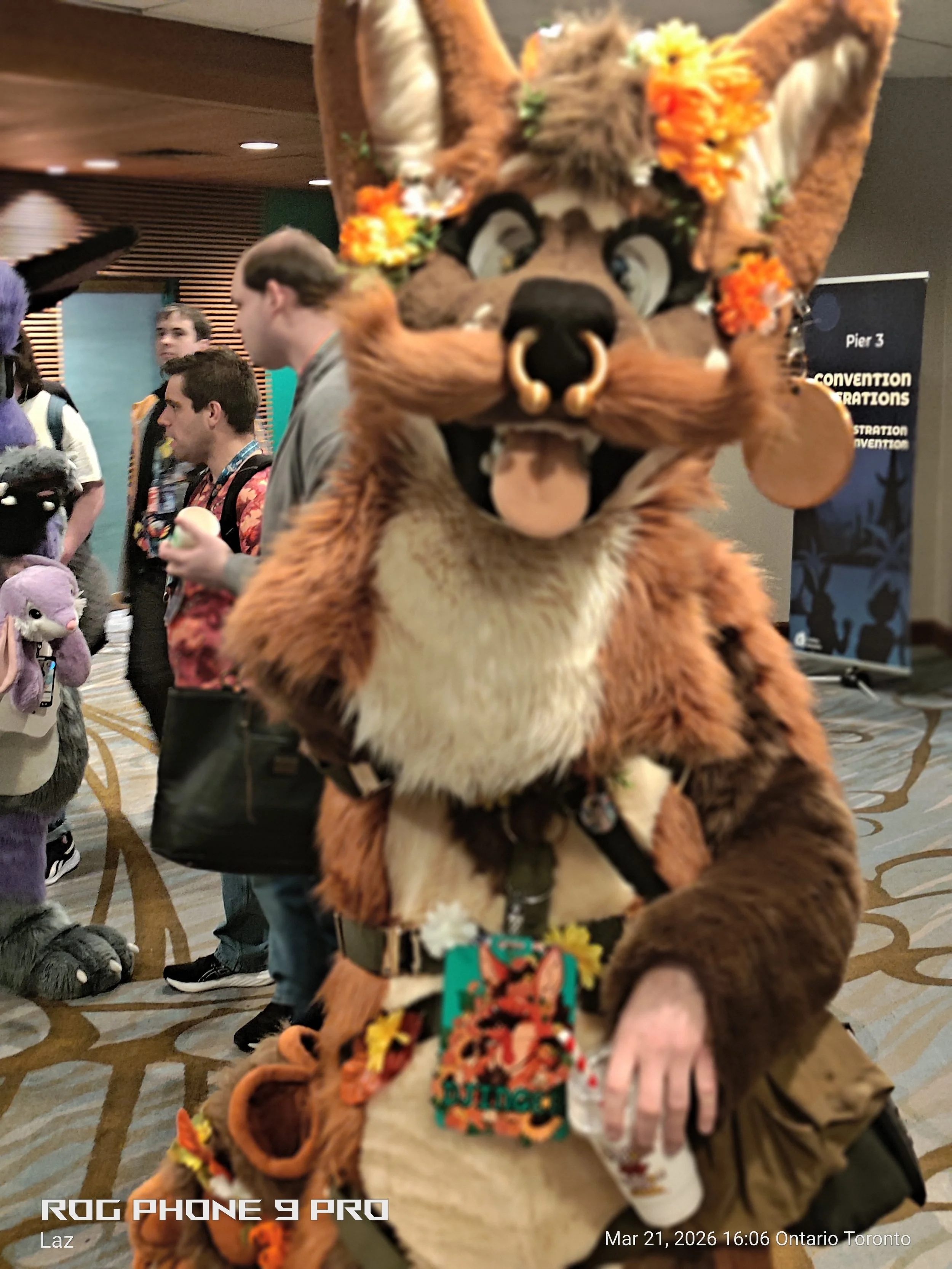 A person dressed in a lion costume with flowers on the head, holding a drink, at a convention or event. Several people and stuffed animals are visible in the background.