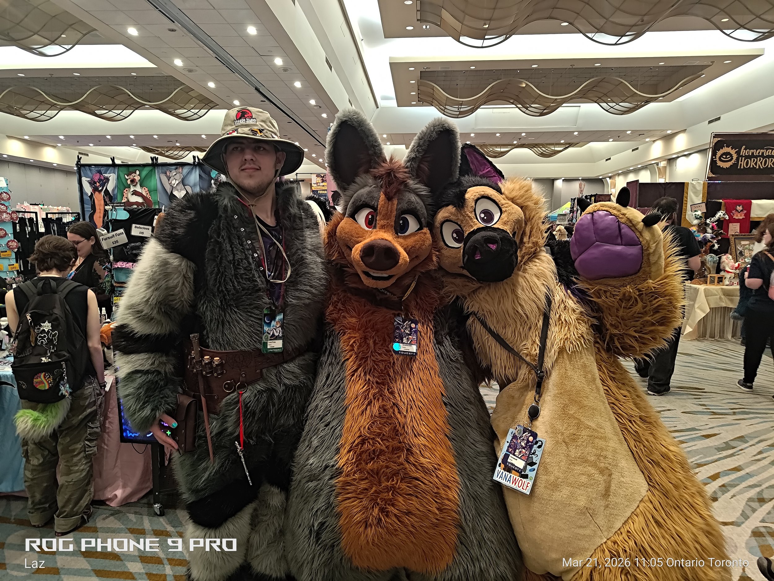 Three people in fursuits posing at a convention, with a person in a naturalistic animal costume and two other people in colorful costumes. The background shows booth displays and other attendees.
