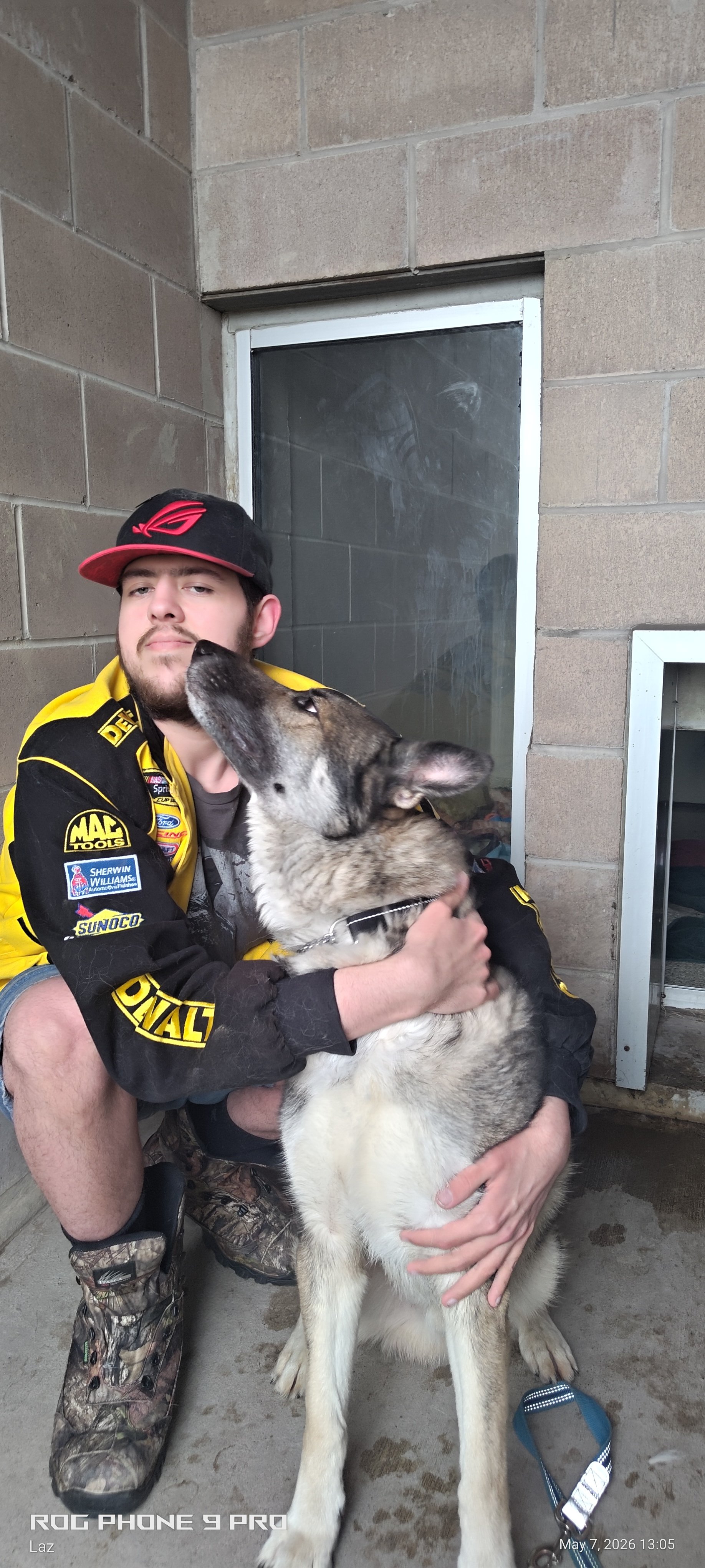 A young man in a racing jacket and camo boots sitting on the ground, smiling and holding a large husky dog. The dog is giving him a kiss on the cheek. They are in a room with brick walls and a black window behind them.