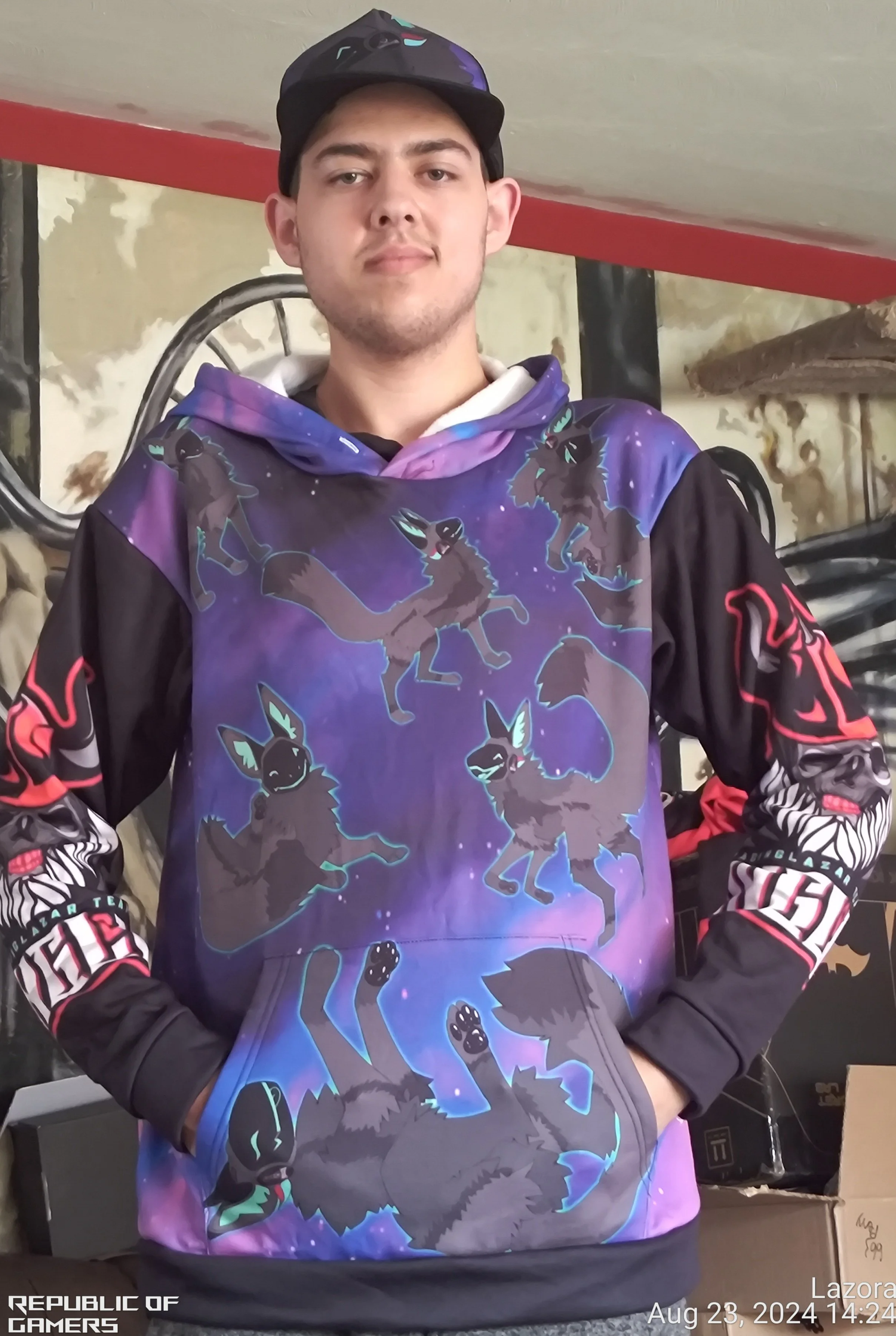 A young man in a colorful hoodie and black cap standing indoors, with a background of a wall, a mirror, and some boxes.