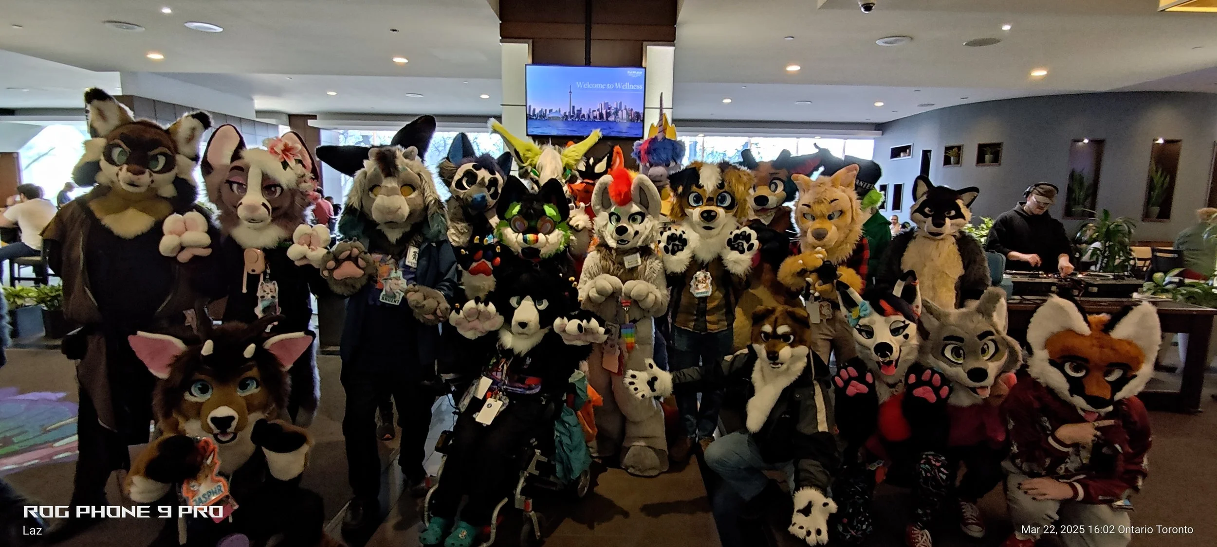 A large group of people dressed in colorful animal costumes, called fursuits, posing together indoors at a convention or event with a sign in the background that says "Welcome to Wellness."