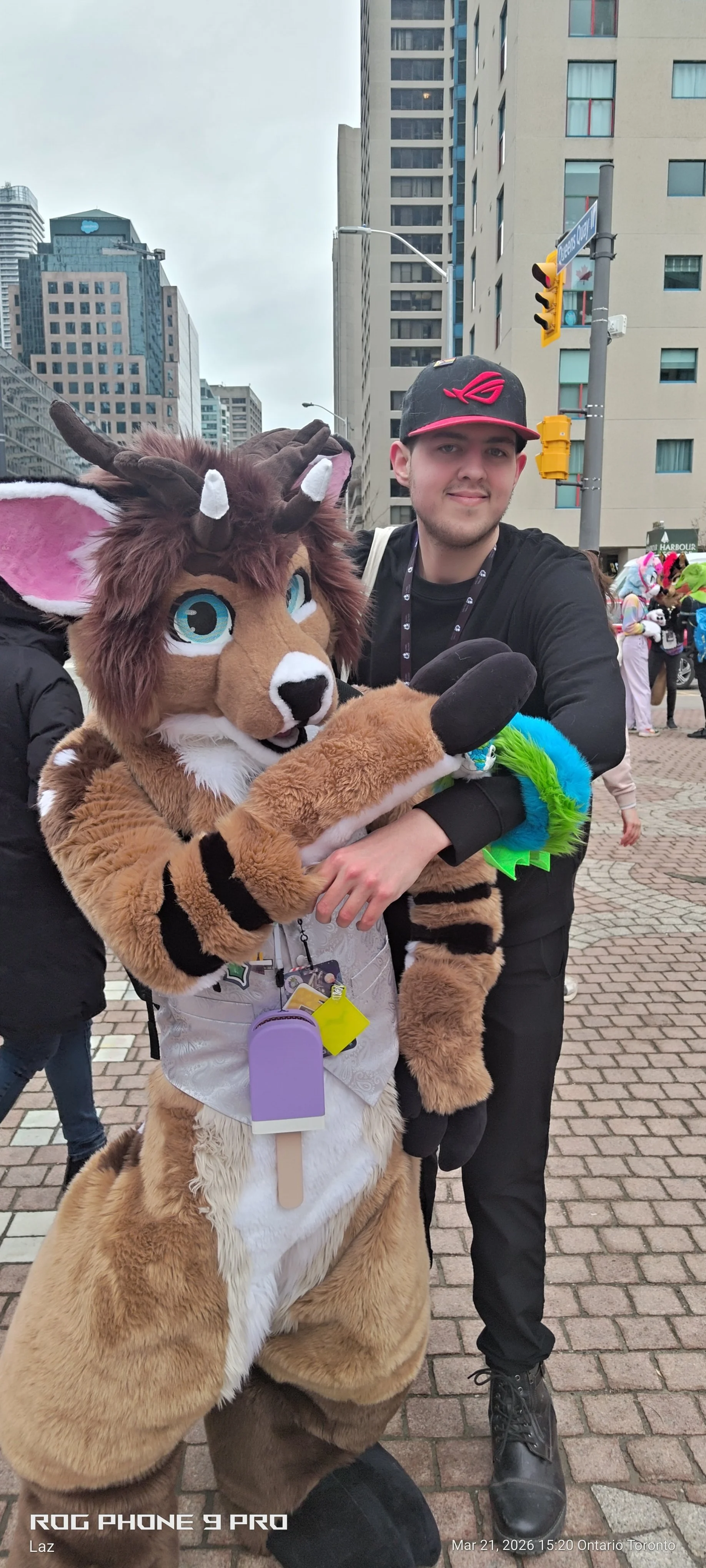 A person in a lion costume and a man in casual clothing are hugging in a city street with tall buildings and a crowd, some of whom are in colorful costumes, in the background.