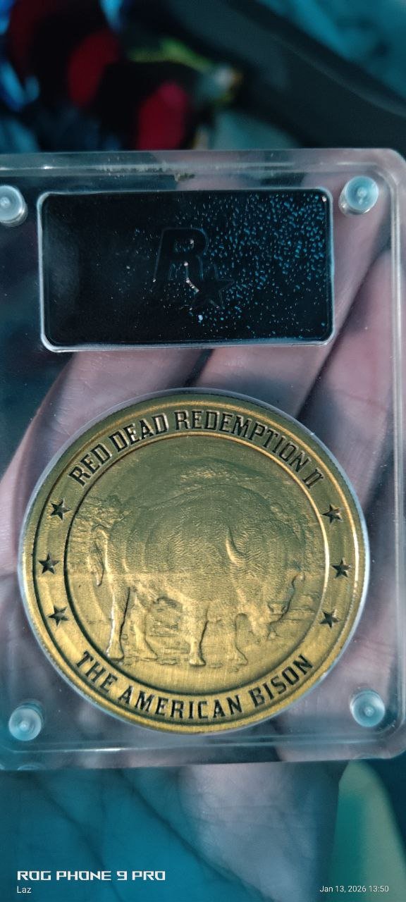 A collectible gold-colored coin inside a plastic display case. The coin is labeled "Red Dead Redemption II" and "The American Bison", featuring an embossed image of a bison.