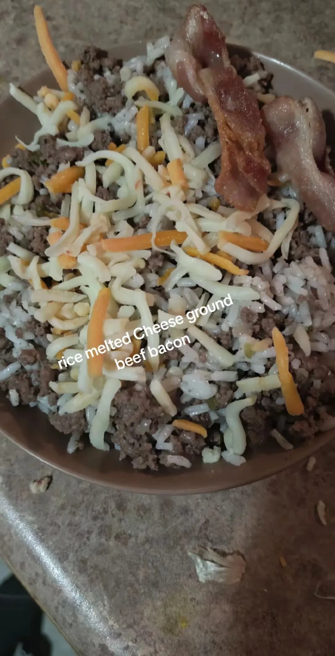 A bowl of rice topped with melted shredded cheese, cooked ground beef, and bacon strips.