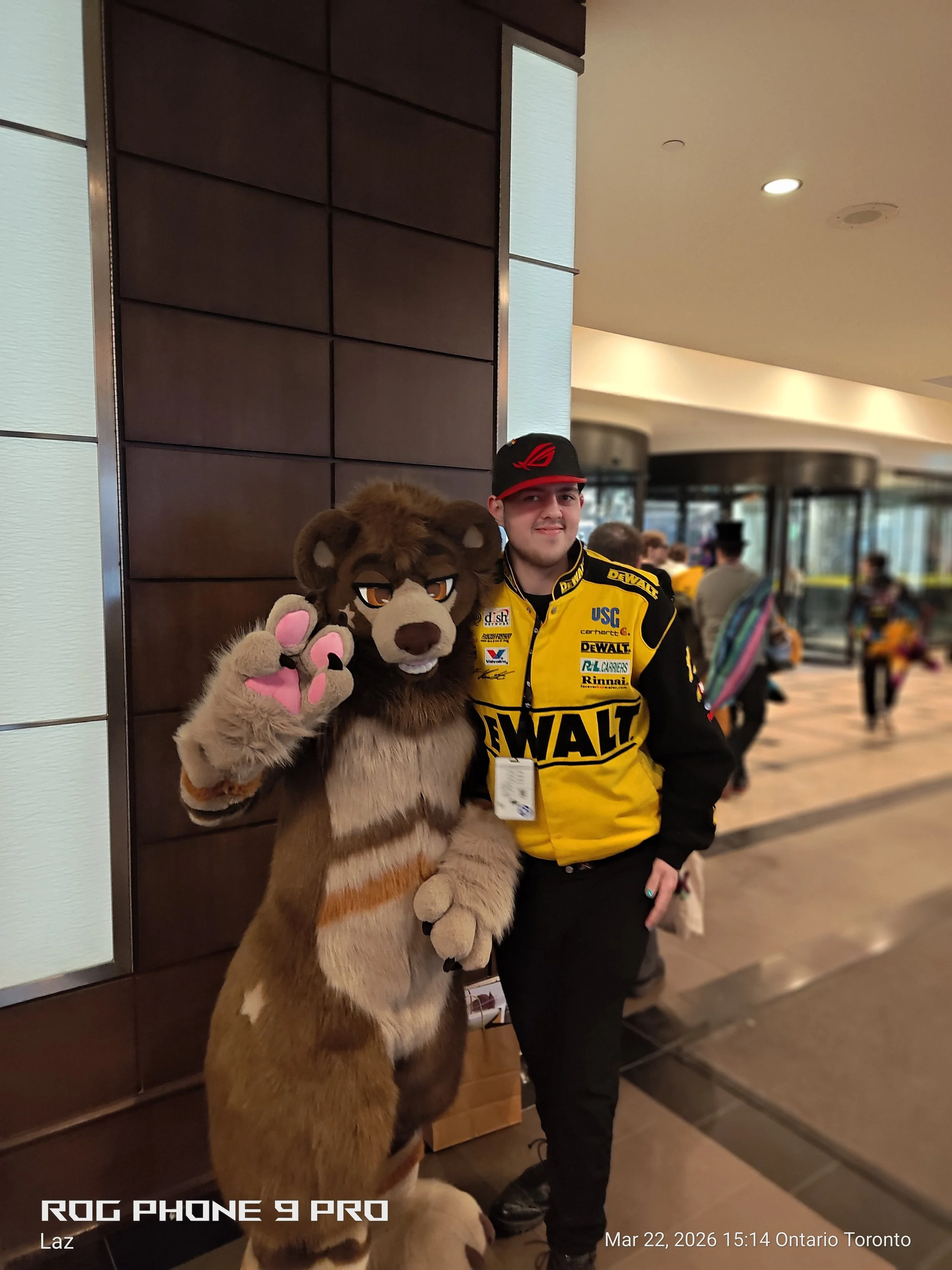 A person dressed in a tiger mascot costume standing next to a man wearing a yellow racing jacket and a black cap, inside a building with a busy background of people and glass doors.