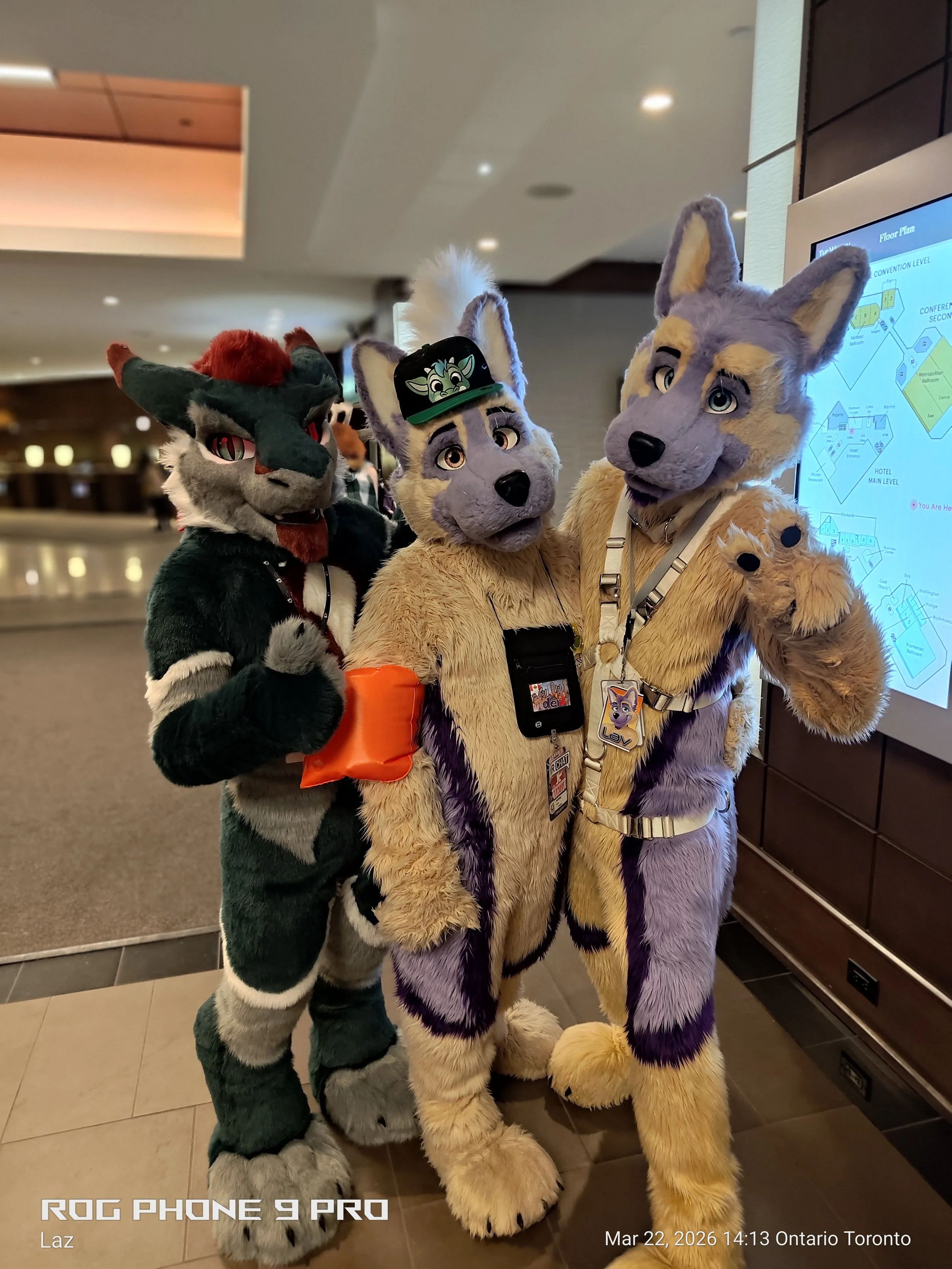 Three people dressed in colorful fursuits of fantasy animals standing in a lobby area. One has black and grey fur with red hair and red eyes, another has purple and cream fur with large ears and a cap with a cartoon character, and the third has yello