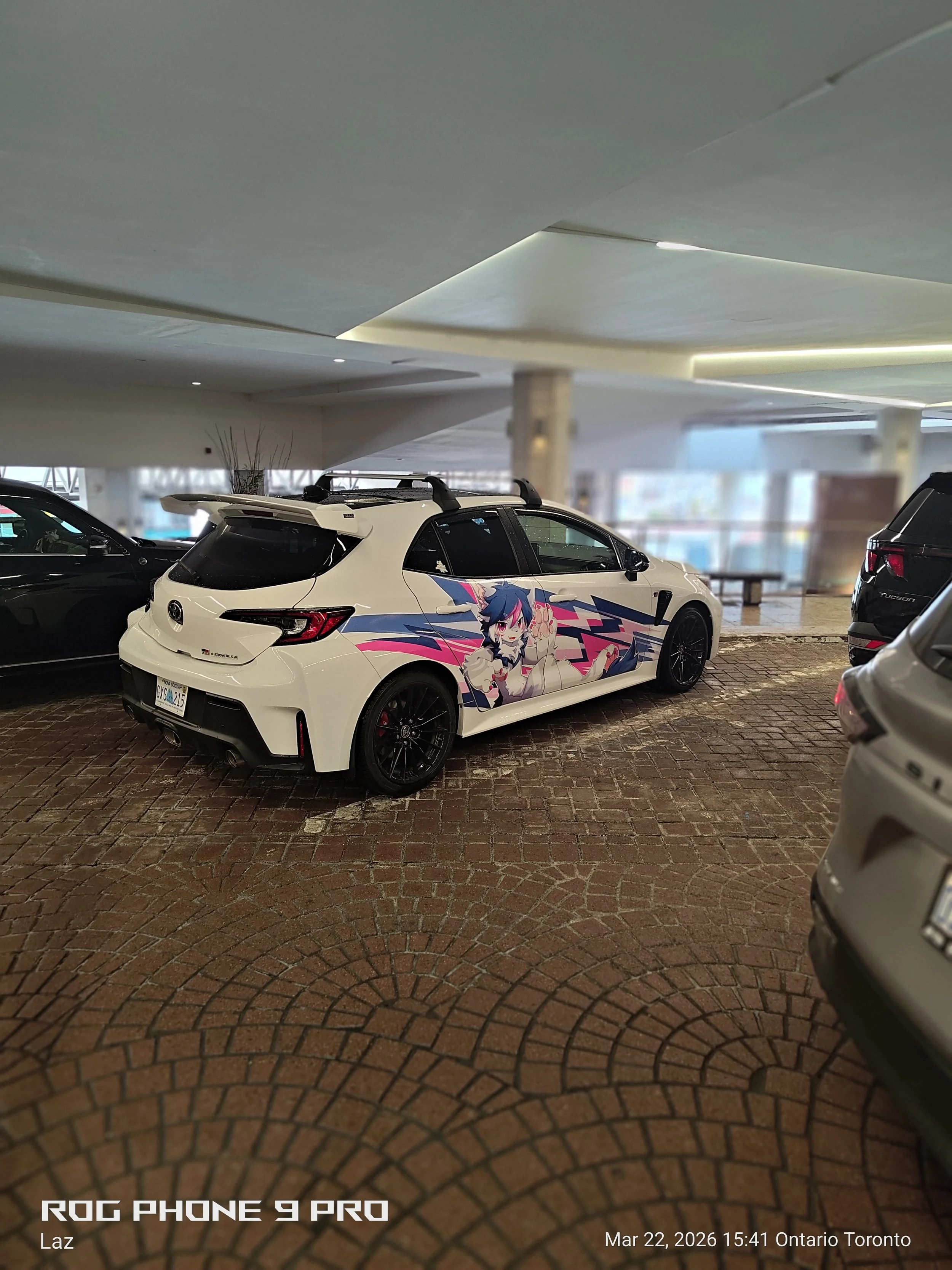 A white car with anime-style character artwork on its side is parked in an indoor parking garage, surrounded by other vehicles, with a modern ceiling design and brick flooring.