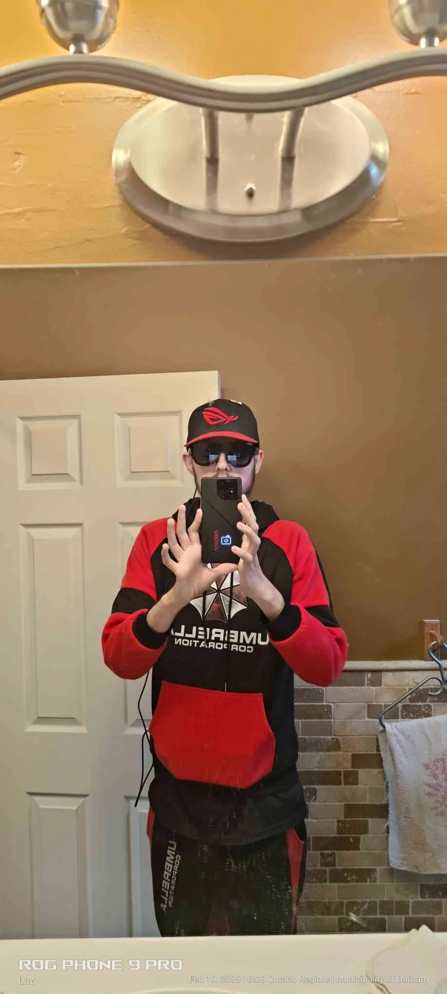 Self-portrait of a person wearing sunglasses, a black and red hoodie with the words "University of" and "Cor..." on it, and headphones, taking a photo in a bathroom mirror. The person is holding a phone, and the bathroom has a door, a towel hanging on a rack, and brick-patterned wall tiles.