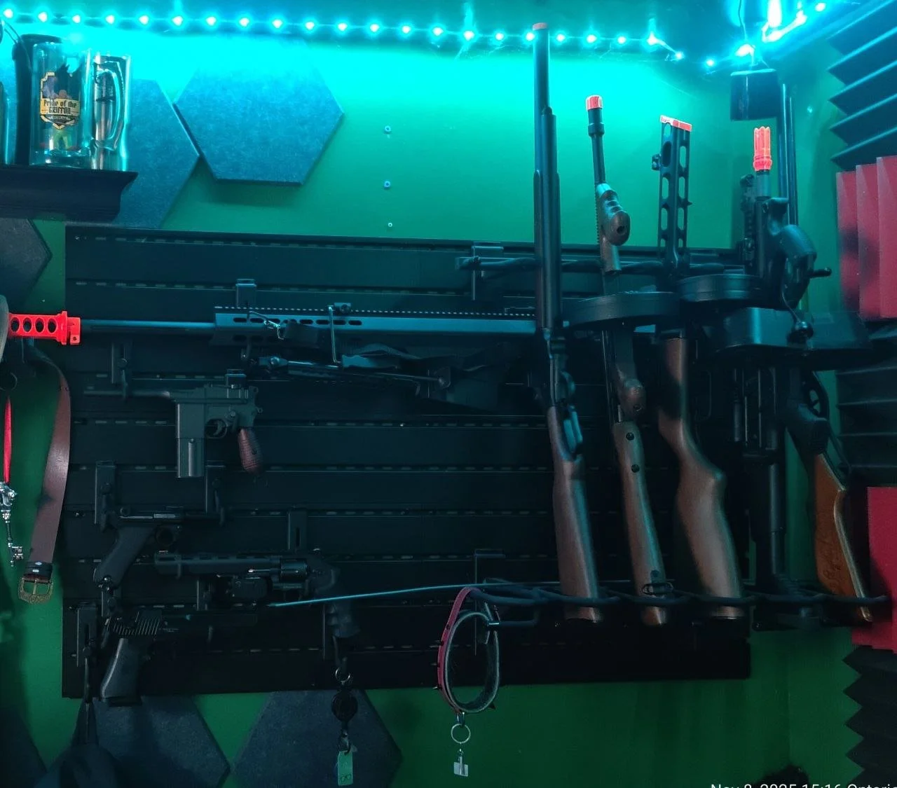 Display case with three airsoft guns, some accessories, and a mug on the top shelf, all against a green wall with soundproofing foam panels.
