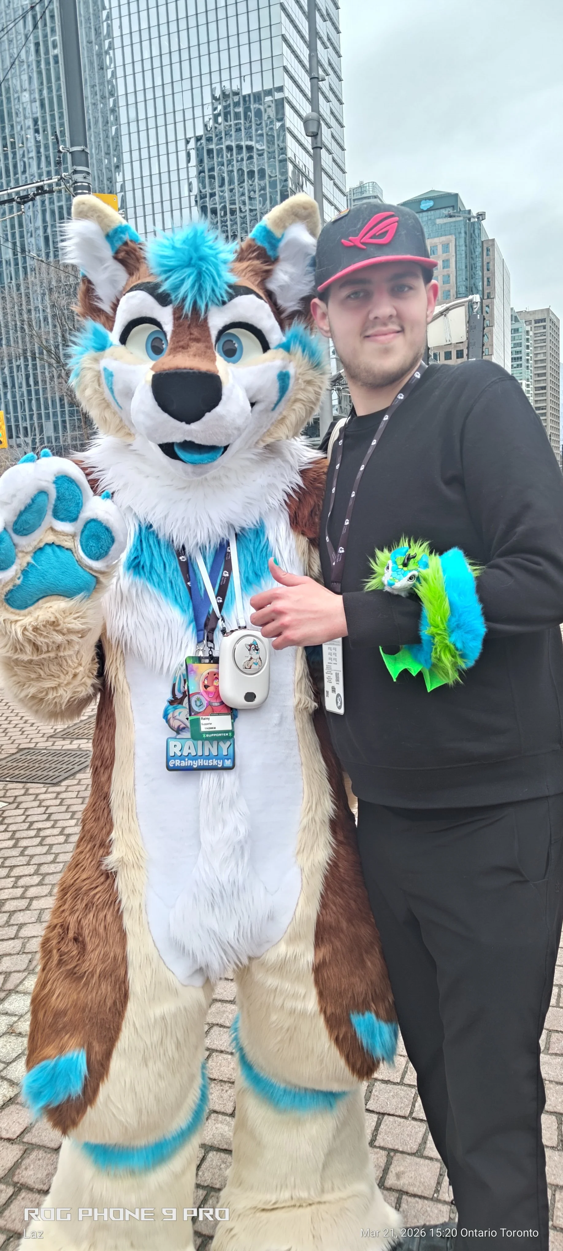 A person dressed in a black outfit and wearing an ROG cap standing next to a colorful anthropomorphic fursuit character with blue, white, and tan fur, in an urban setting with tall glass buildings.