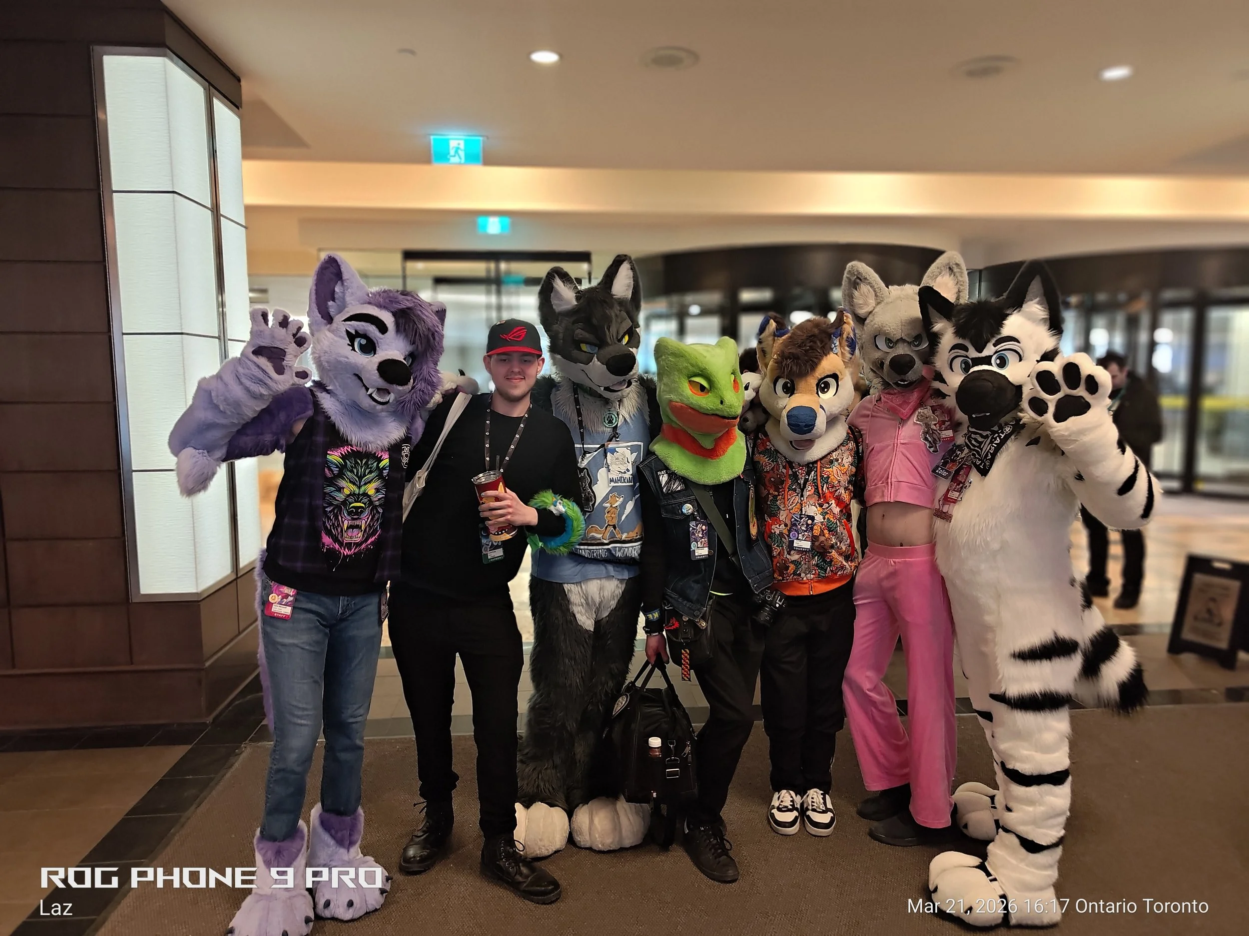 Group of people dressed in colorful animal fursuits and costumes, standing together indoors at an event, with some making gestures and smiling for the camera.