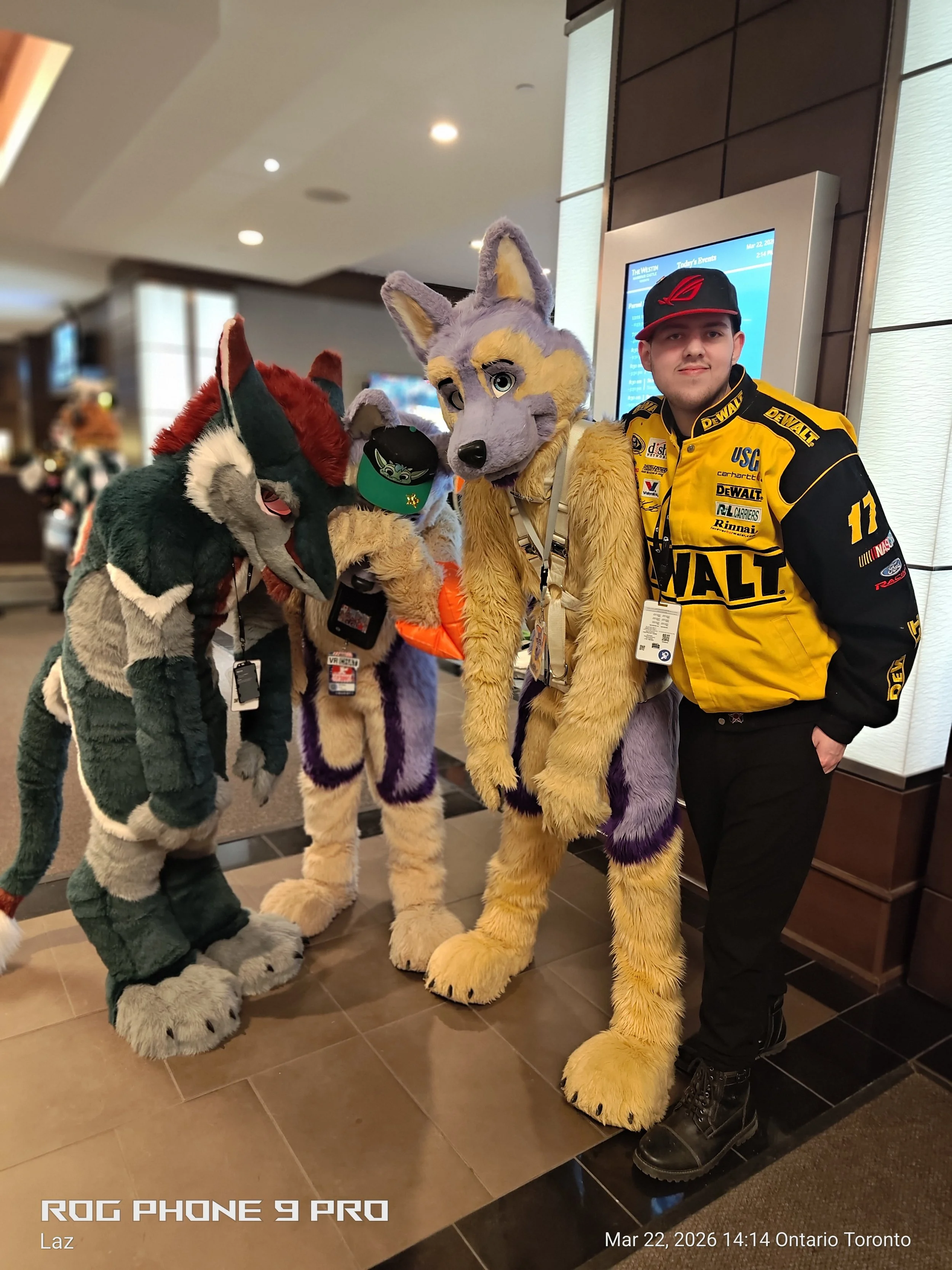 A man in a yellow and black racing jacket and black pants is standing next to three people in animal costumes, including a wolf, a fox, and a smaller fox, in an indoor setting.