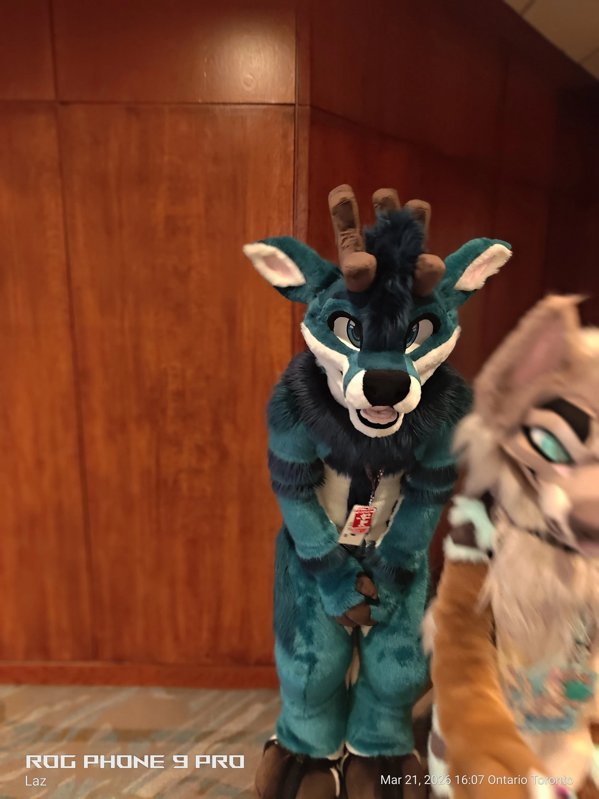 A plush toy resembling a blue and black wolf or fox with antlers, large eyes, and a friendly expression, standing upright with a wooden background behind it.