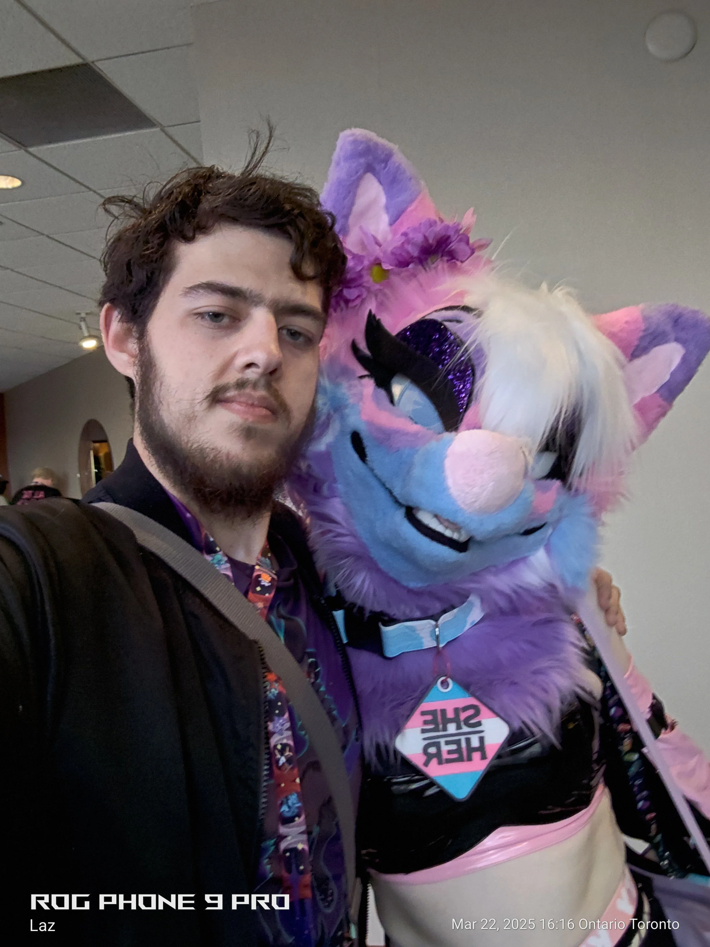 A young man with a beard and curly hair taking a selfie with a person in a colorful unicorn costume at an indoor event, with a ceiling and other people in the background.
