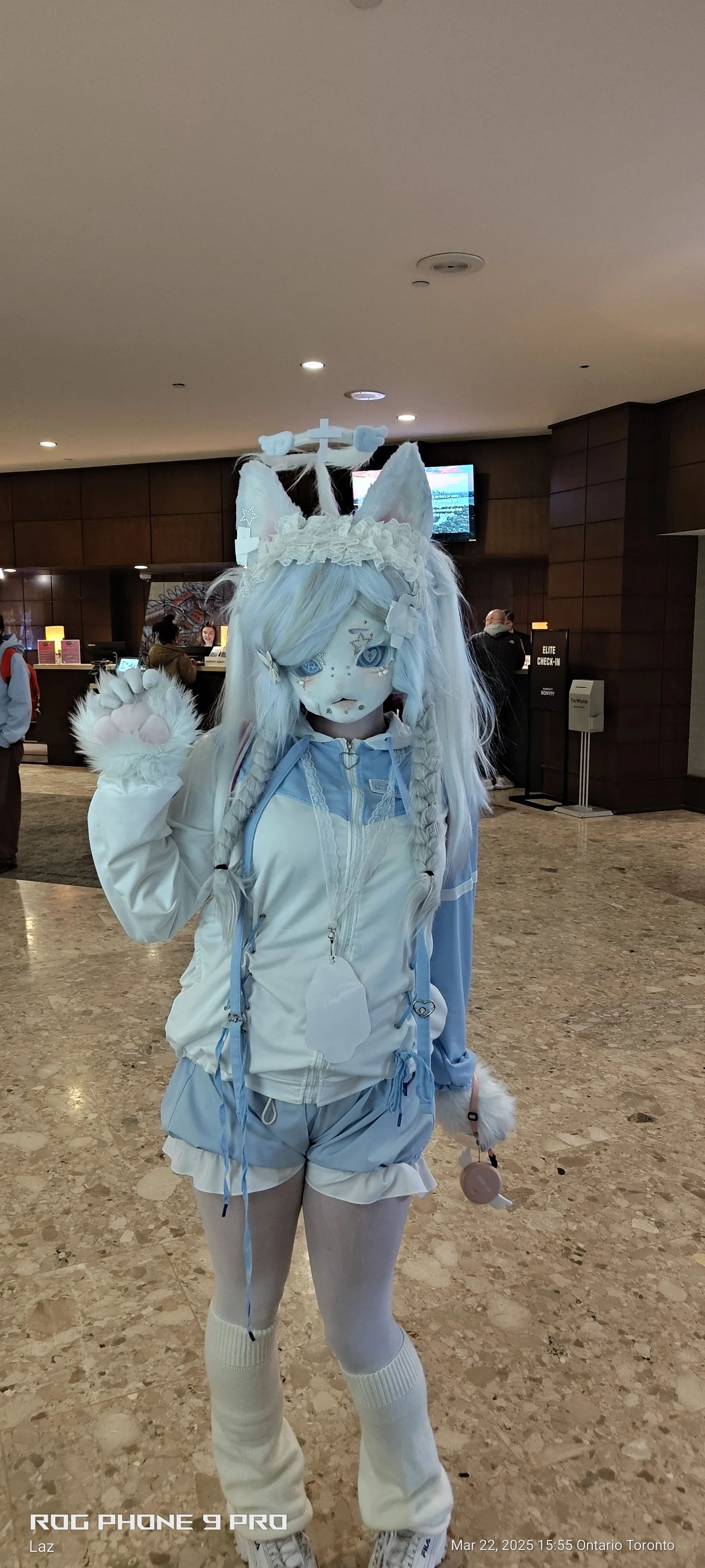A person dressed in a white and blue animal-themed cosplay costume with a tail, cat ears, and detailed face makeup, standing indoors at a lobby or reception area.