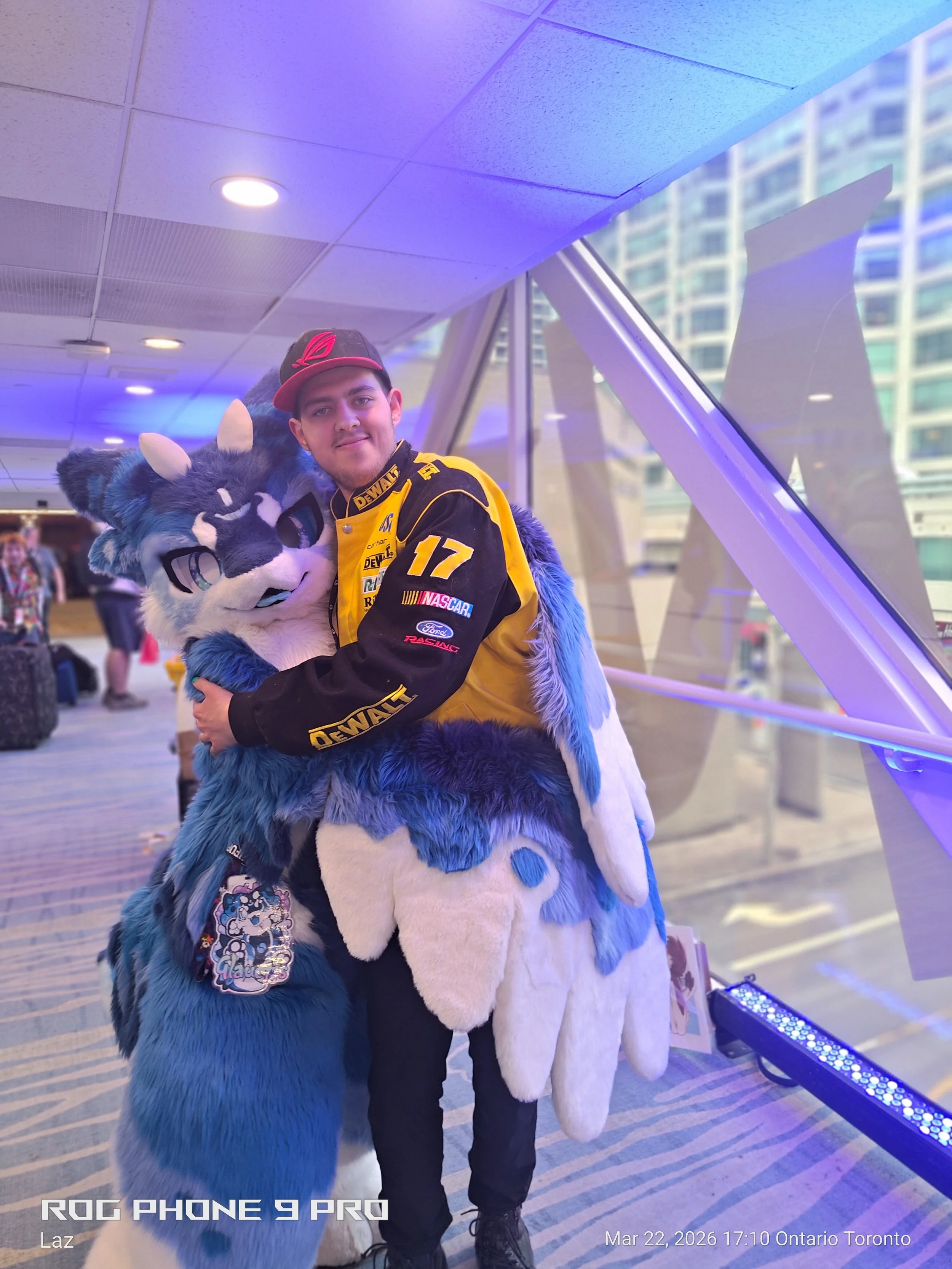 A person in a yellow and black racing jacket hugging a blue and white person in a fursuit of a wolf or fox. They are in an indoor space with a glass wall and city buildings visible outside.