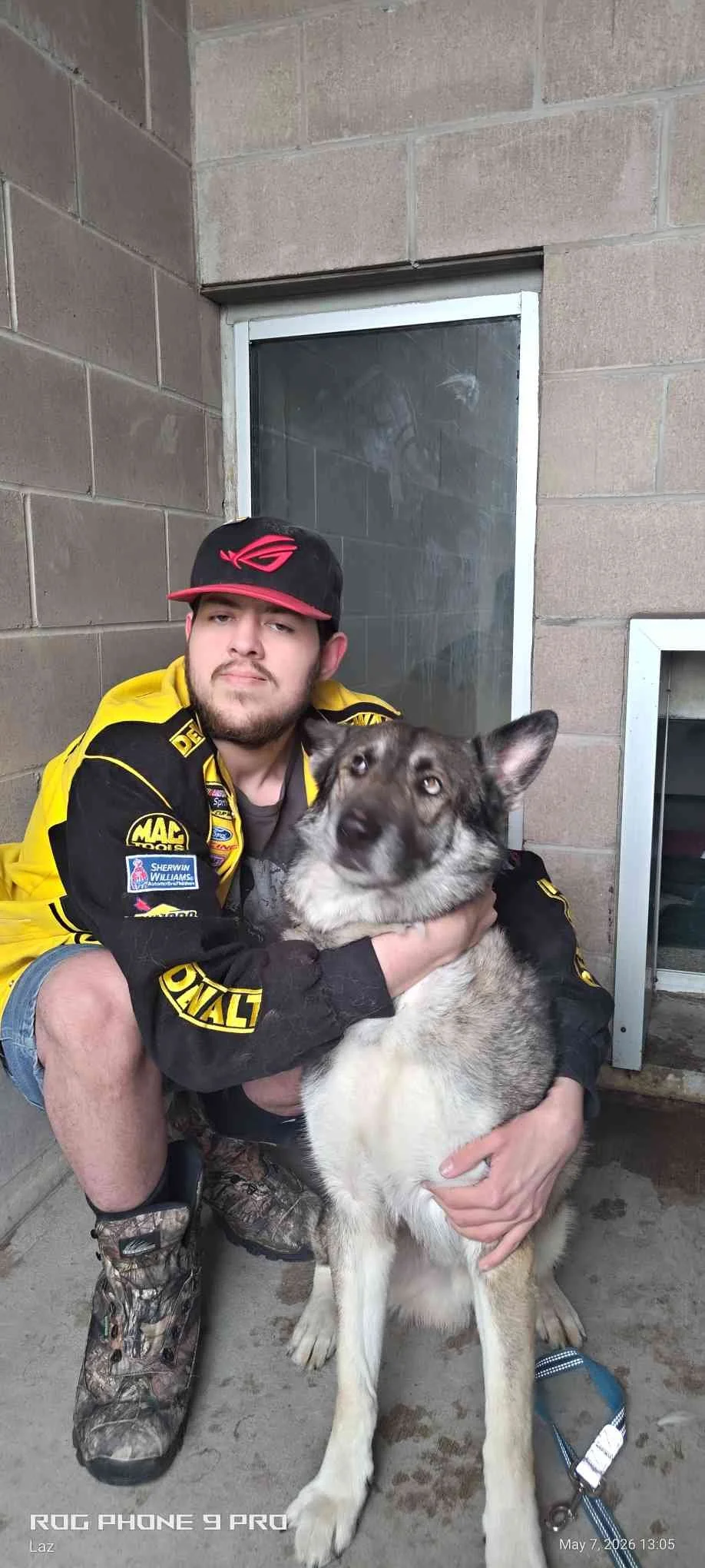 A young man wearing a racing jacket and cap crouches beside a large gray and white Siberian Husky dog, holding its neck and chest. They are outside against a concrete wall with a small window and a vent.