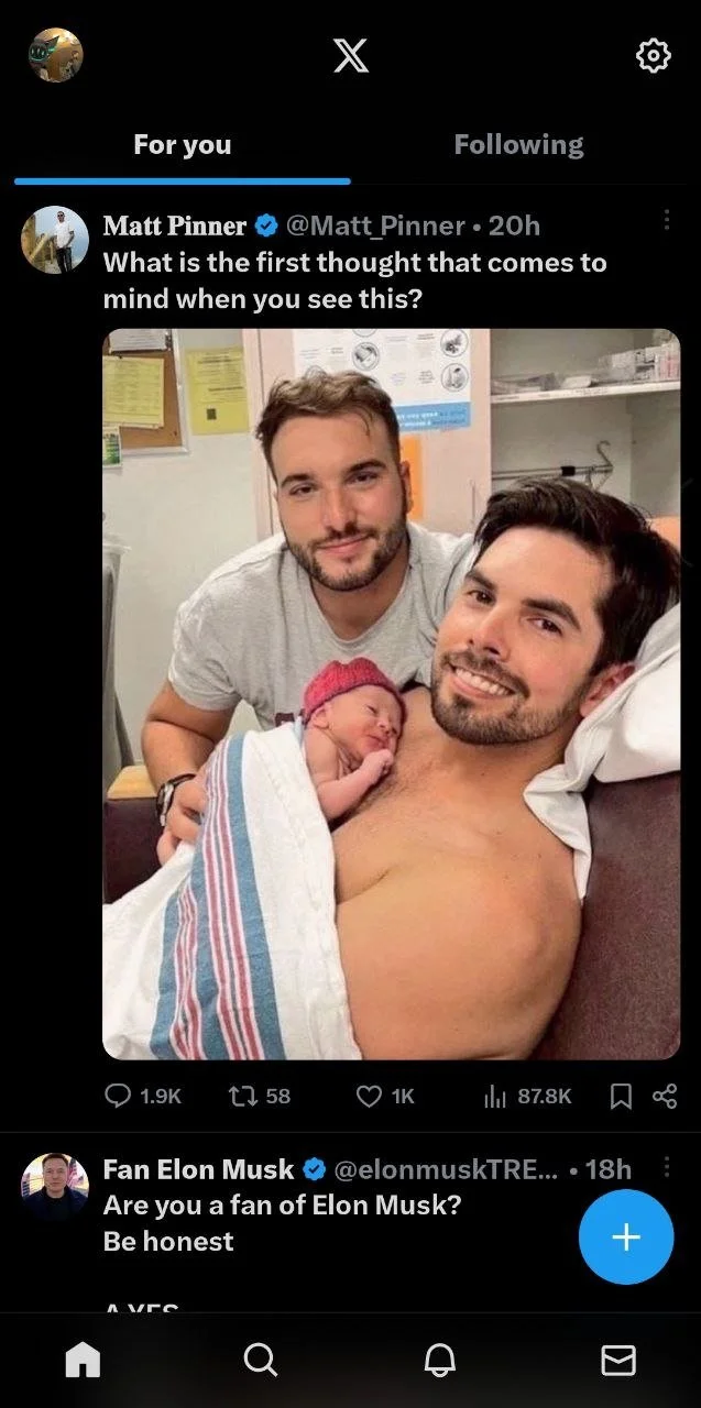A man is lying in a hospital bed with a newborn baby on his chest, wrapped in a hospital blanket, and another man is standing beside them. The scene appears to be in a hospital room.