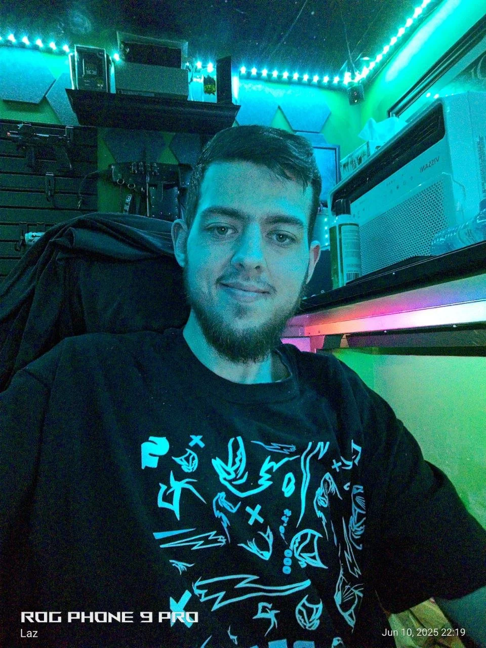 A young man with dark hair and a beard taking a selfie in a colorful room with blue and green lights.