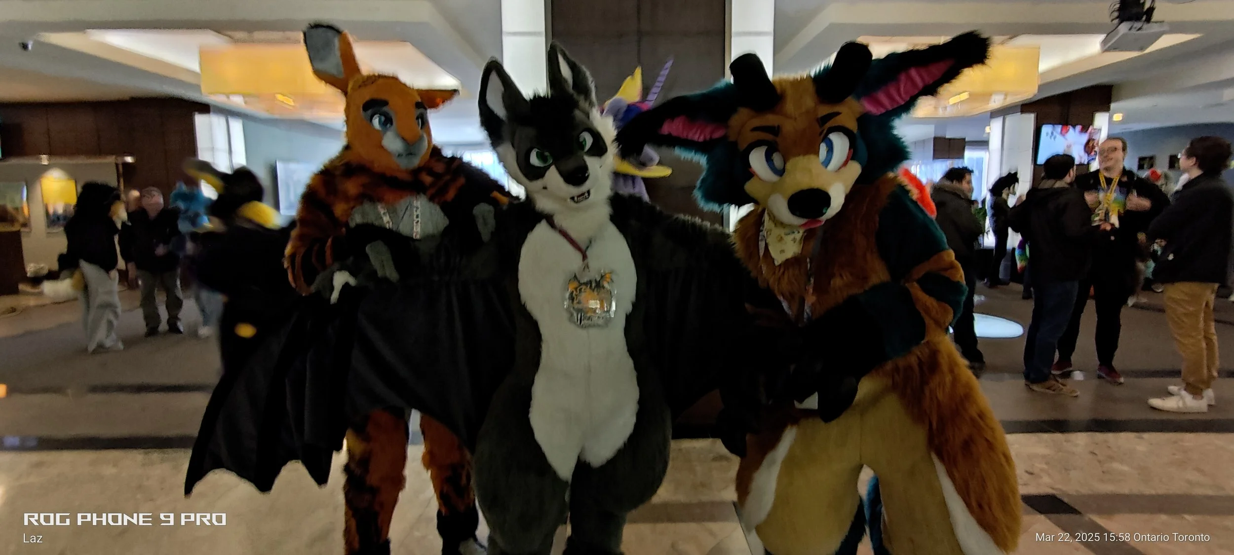Three people dressed in animal costumes, including feline, fox, and canine characters, standing inside a lobby area at a furry convention with other attendees in the background.