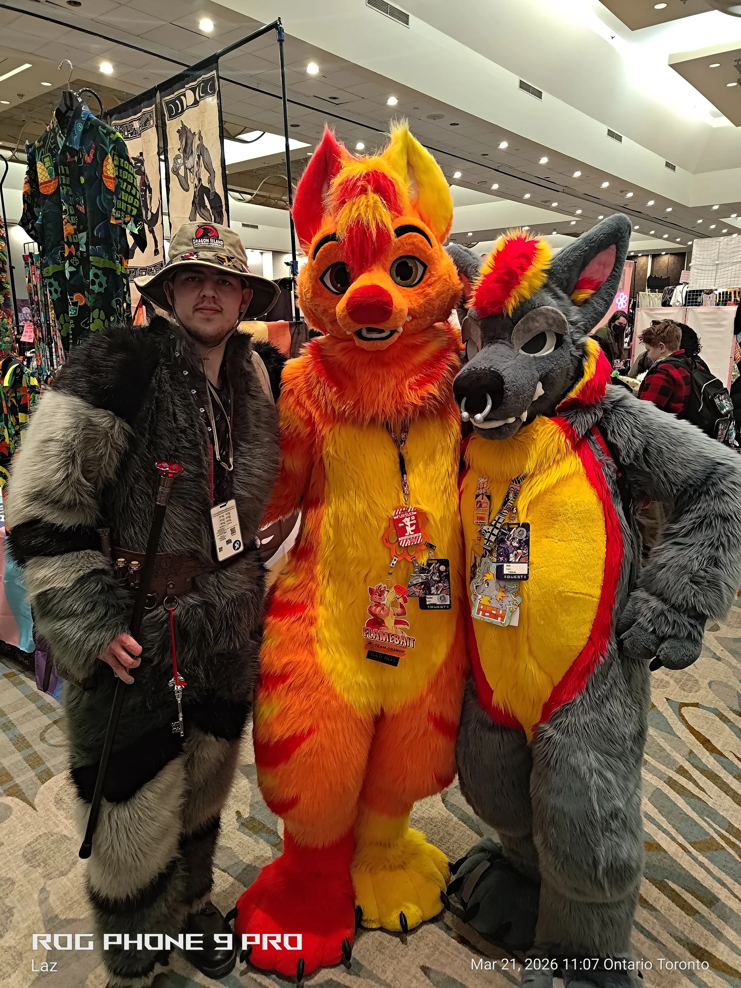 Three people in furry animal costumes, including a bright yellow and orange fox and a gray wolf, standing together at a convention or event in a large indoor space with booths and attendees in the background.