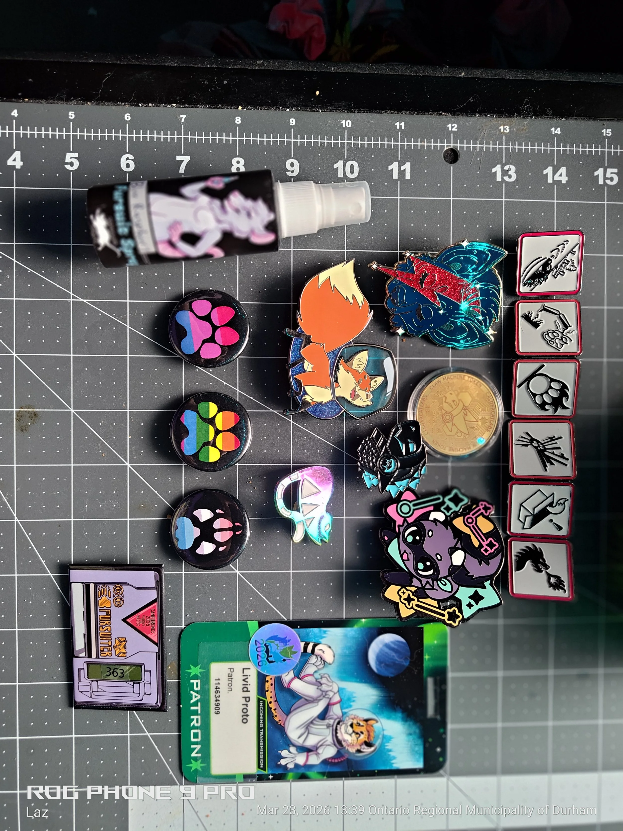 A collection of various items on a black cutting mat with measurement grid, including enamel pins of characters and symbols, a small coin, a colorful badge, a trading card, a sticker, and a spray bottle.