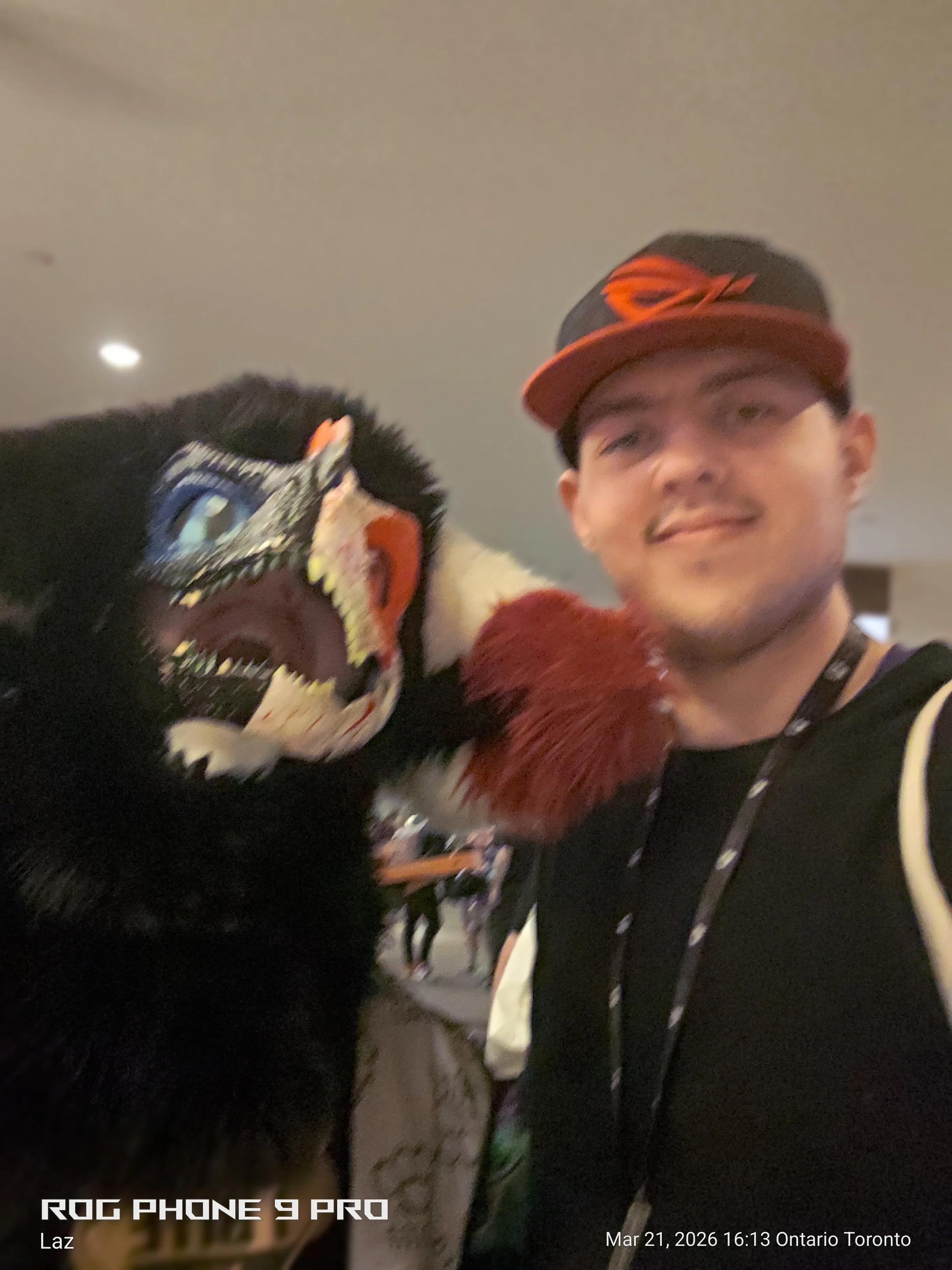 A young man wearing a black and red cap, a black jacket, and a lanyard around his neck taking a selfie with a person dressed as a black panther mascot.