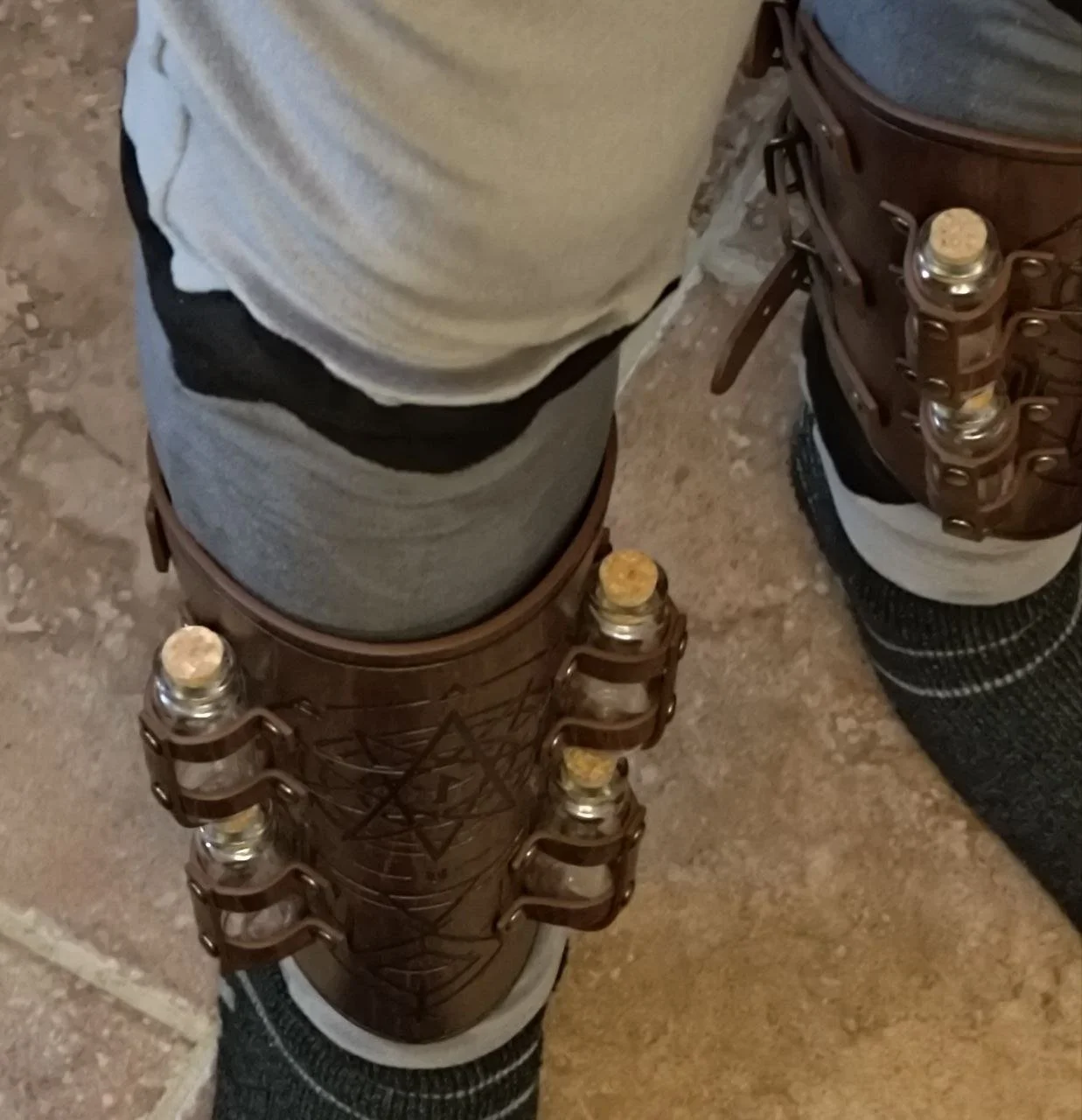 Person standing on a tiled floor wearing dark boots, gray pants, a gray and black hoodie, and brown leather ankle holsters with metal details.