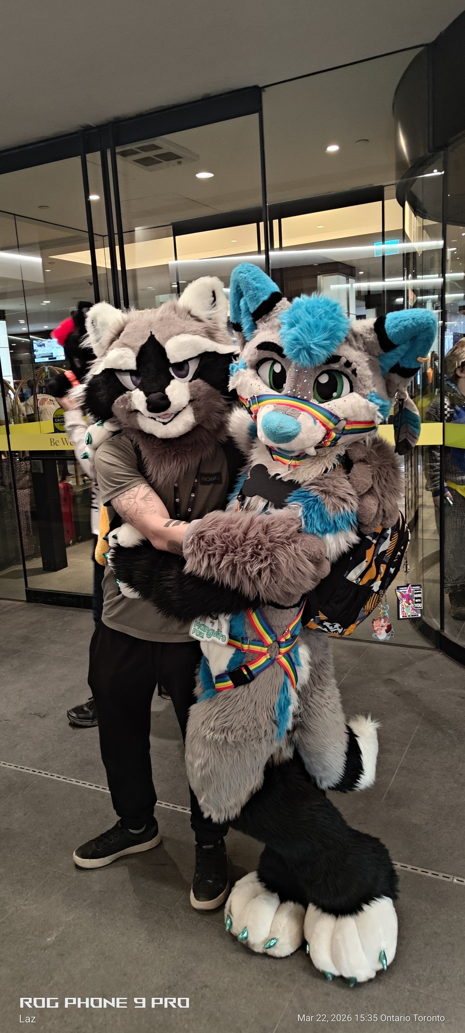 Two people in furry animal costumes hugging in front of a glass door at an airport.