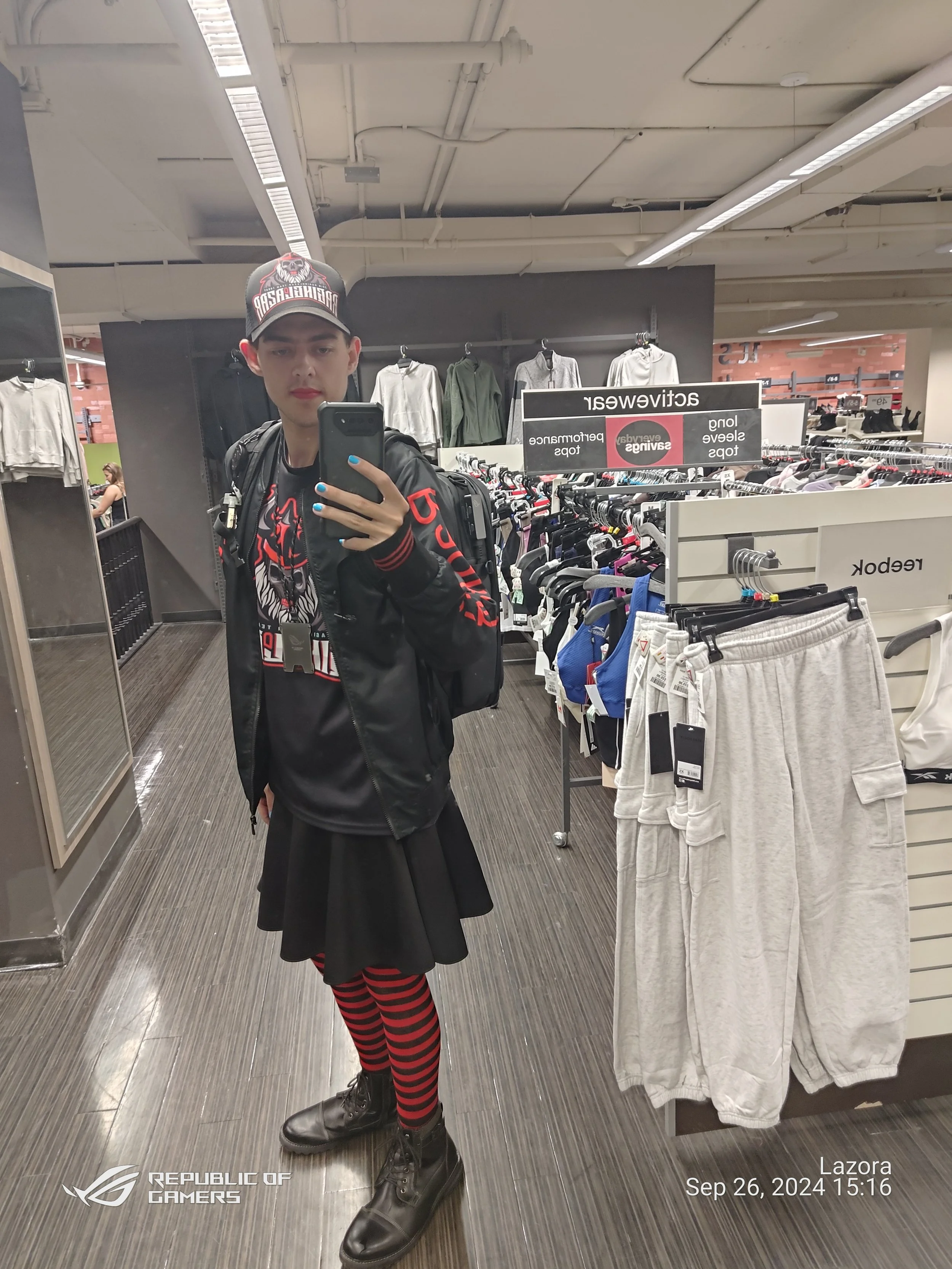 Young man taking a selfie in a store mirror, wearing a black jacket, black shirt, red and black striped tights, black combat boots, a black cap with a graphic print, and carrying a backpack. The store has clothing racks and signs indicating sale or c
