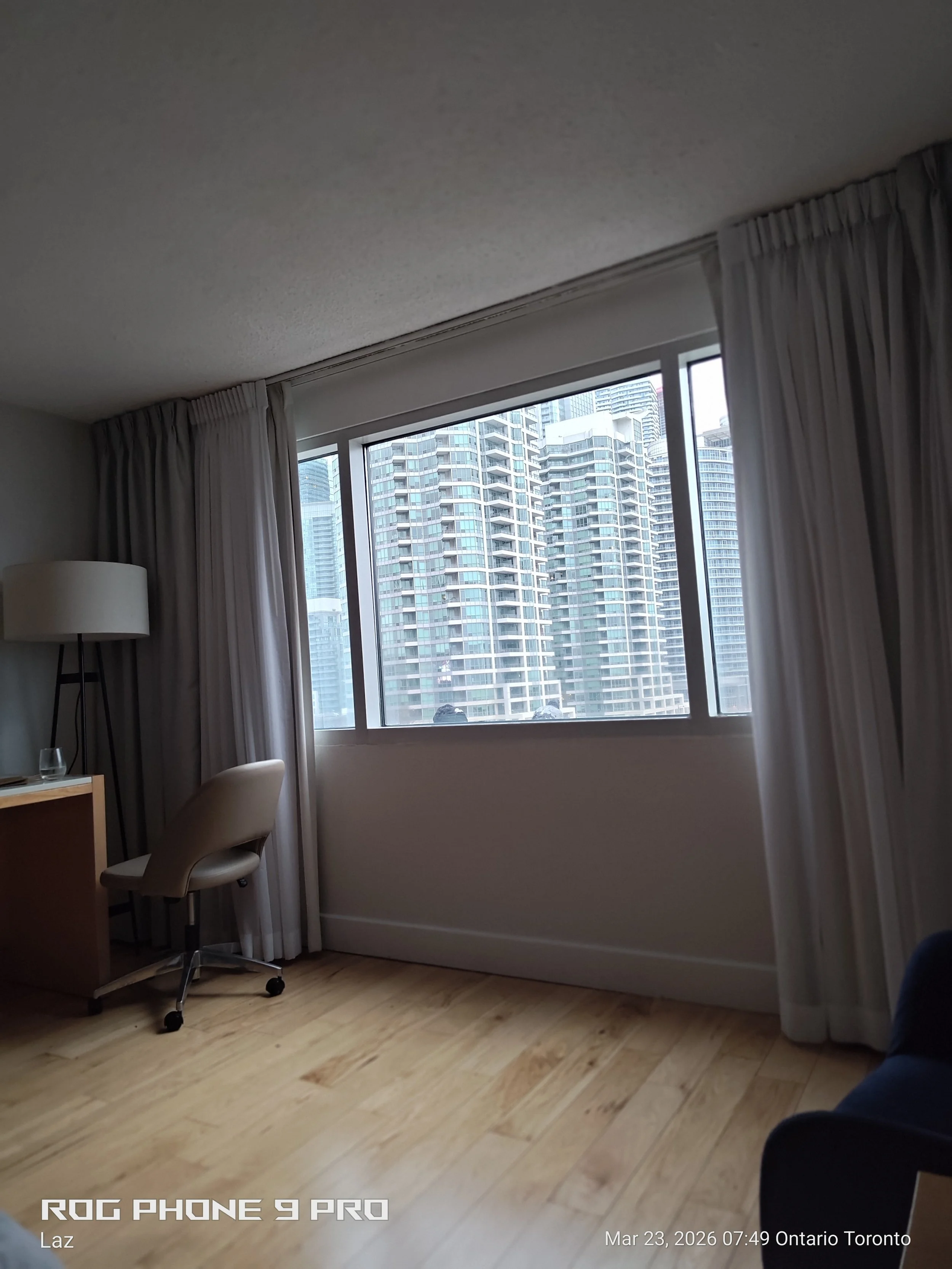 Interior of a room with a large window showing high-rise buildings outside, beige curtains, a beige office chair, a table with a glass, a floor lamp, and light-colored wooden flooring.