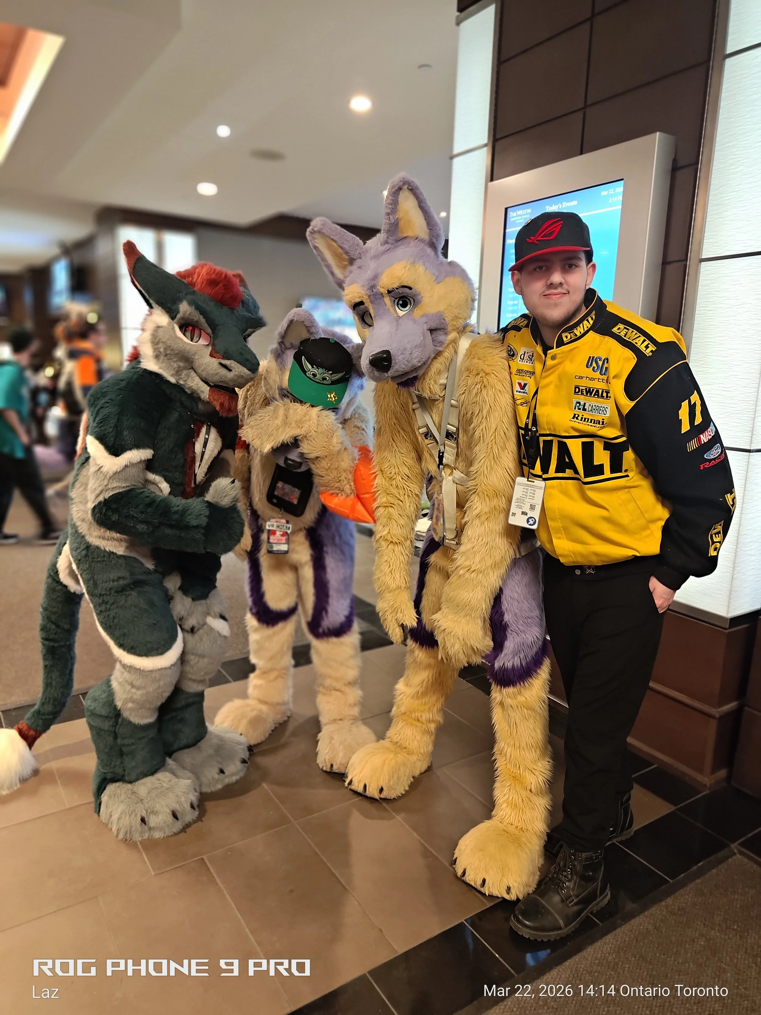A man wearing a yellow and black racing jacket and a black cap with red accents stands next to three people in colorful furry animal costumes, including a fox, a wolf, and a feline, indoors with a digital directory in the background.