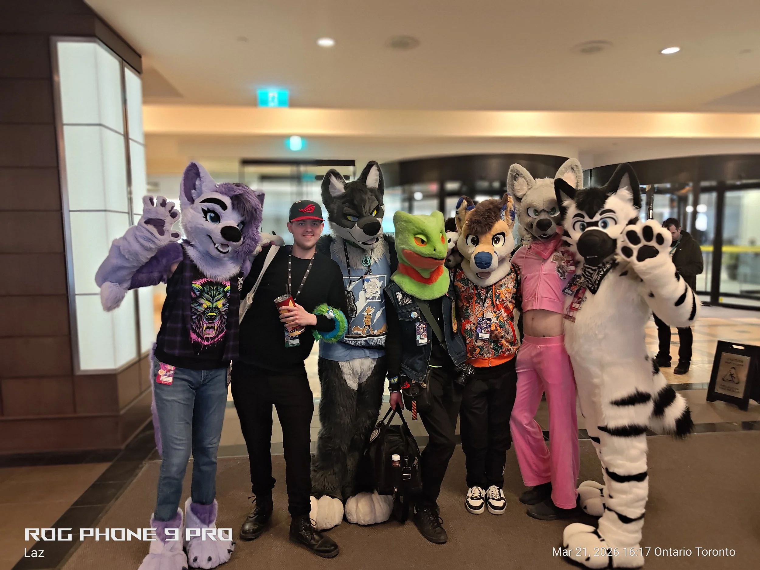 A group of people dressed in colorful animal costumes, including a purple tiger, a gray wolf, a green frog, a beige husky, a black and white husky, and a white tiger, standing together in an indoor convention or event space in Toronto on March 21, 20