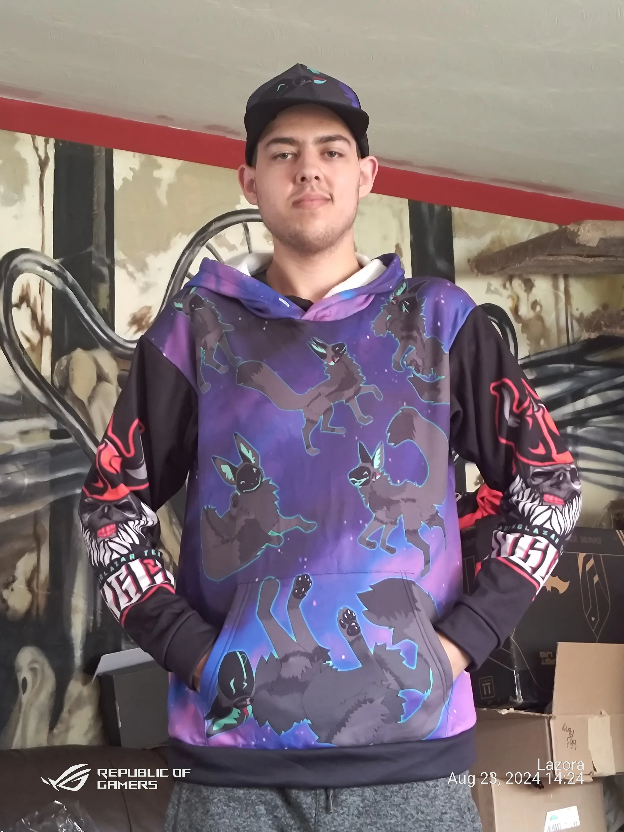 A young man wearing a black cap and a colorful hoodie with a wolf and moon design, standing indoors with a background of stone wall decor and gaming equipment.