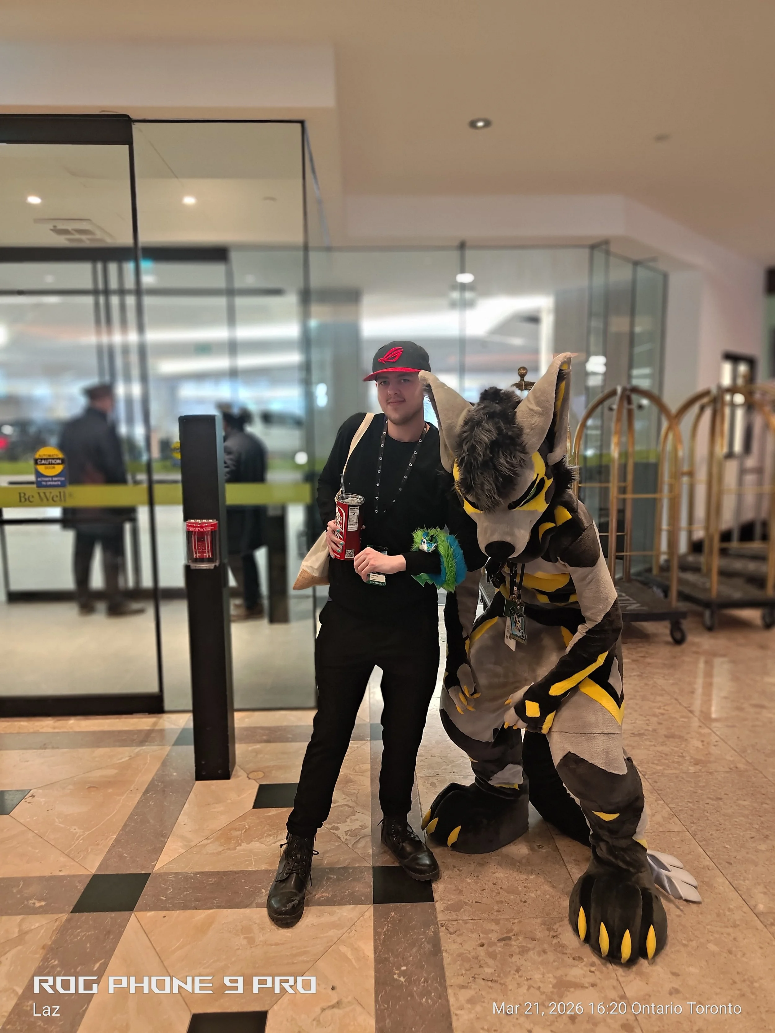 A person dressed in a black outfit and a cap with a red logo stands next to a person in a bee costume at an indoor location, possibly an airport or mall, with glass doors and a tiled floor.