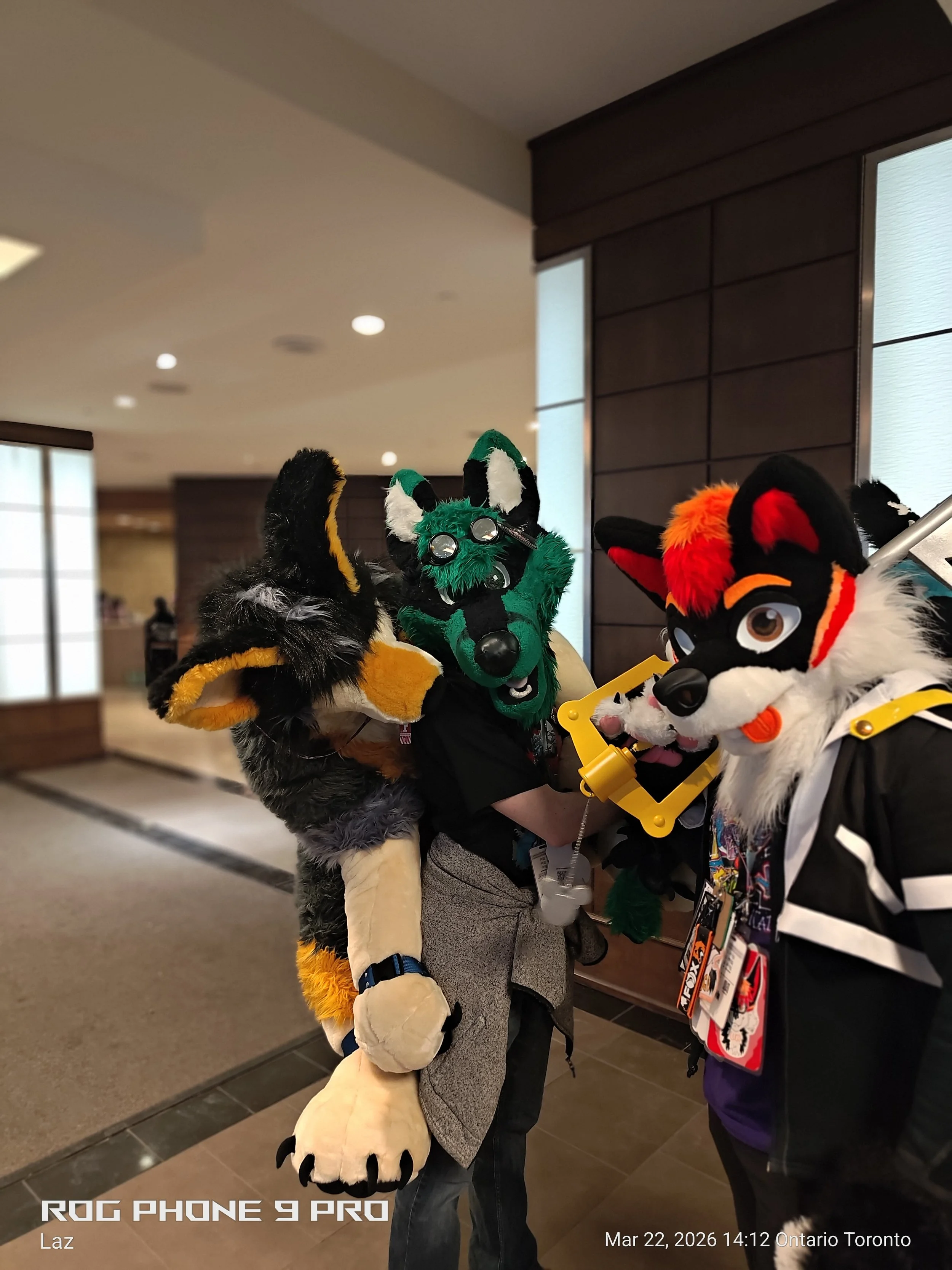 Two people dressed in colorful furry mascot costumes are standing indoors. One mascot is green and black, and the other is black, white, red, and orange. They are close together, engaging in a friendly interaction.