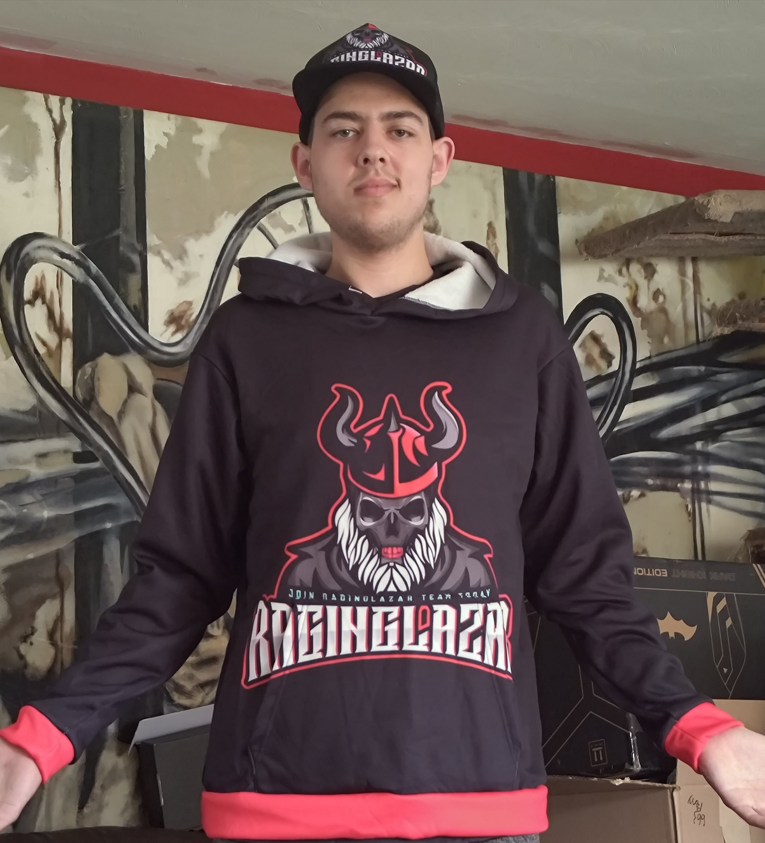 A young man wearing a black hoodie and a black cap with a tiger logo, standing indoors in front of a decorative iron bed frame and a wall with peeling paint. He is facing the camera with a neutral expression.