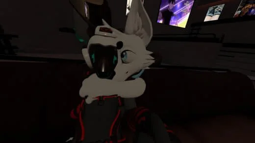Anthropomorphic wolf character with gray and white fur, blue eyes, and black and red clothing, sitting indoors with computer screens in the background.