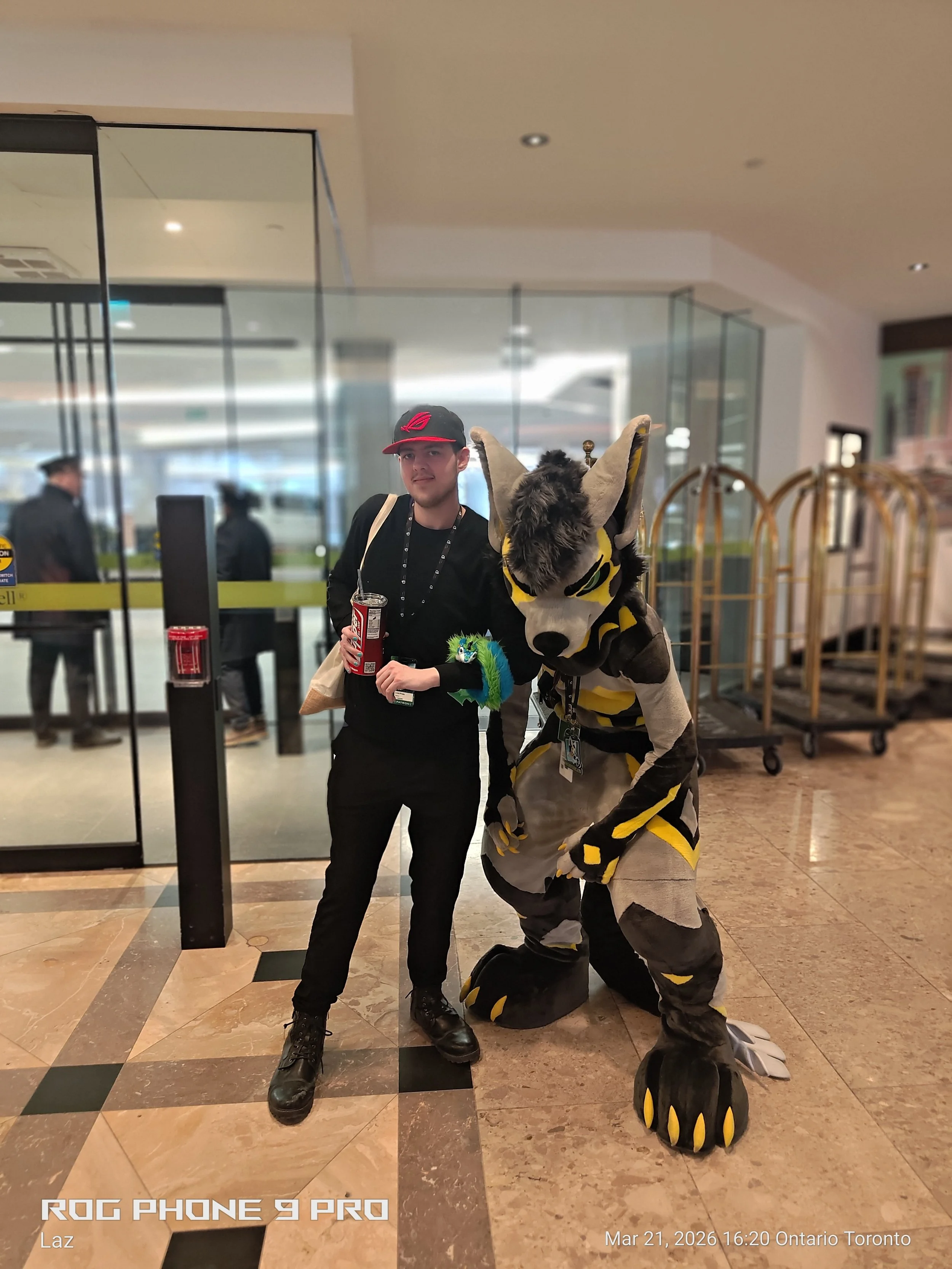 A person dressed in a reptile costume standing next to a young man holding a soda cup inside a building at Ontario Toronto, with luggage carts in the background.