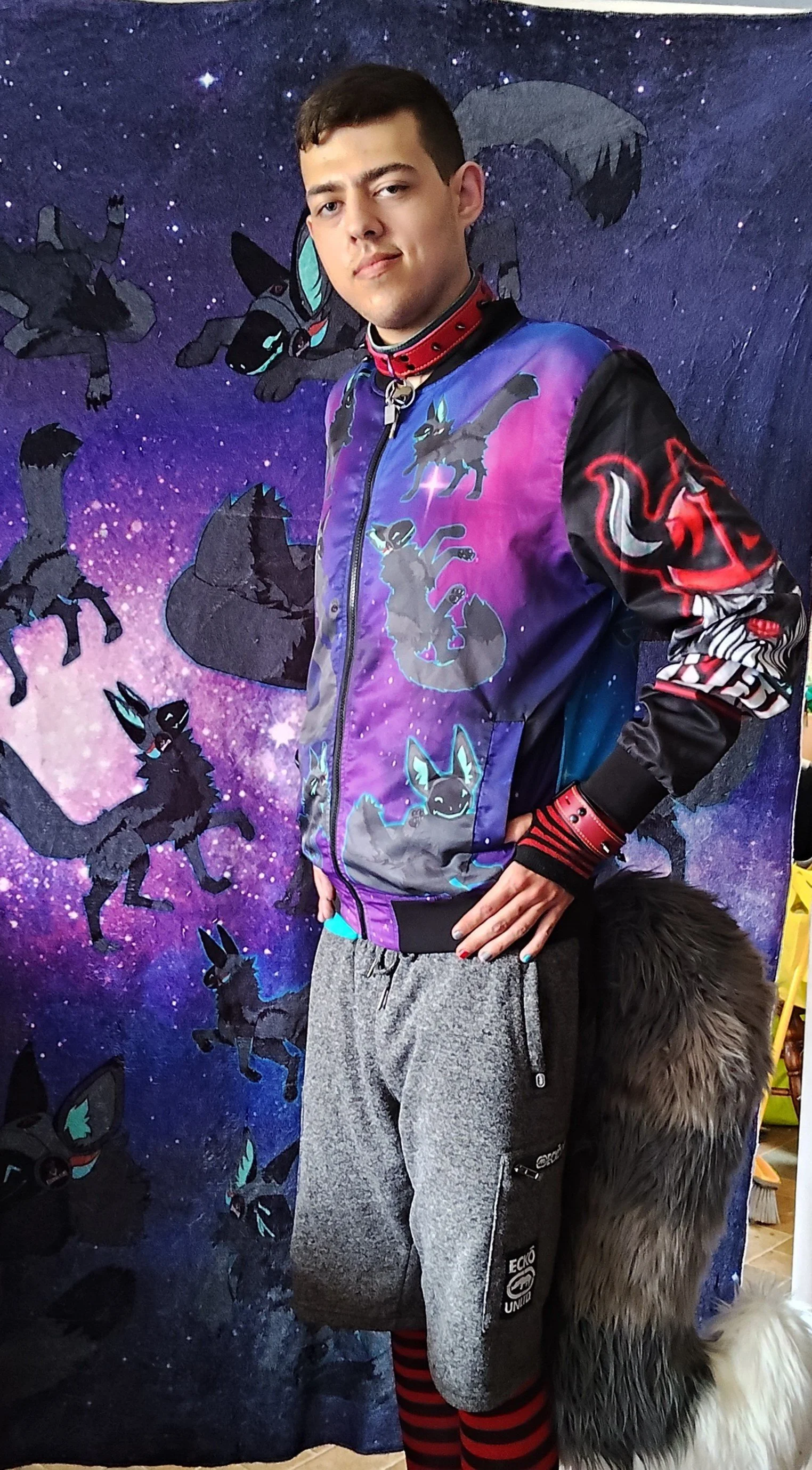 A young man standing in front of a vibrant, space-themed backdrop with cartoon-style flying and standing black fox characters with blue and turquoise markings. He is wearing a colorful jacket with fox illustrations and a red choker, gray sweatpants, 