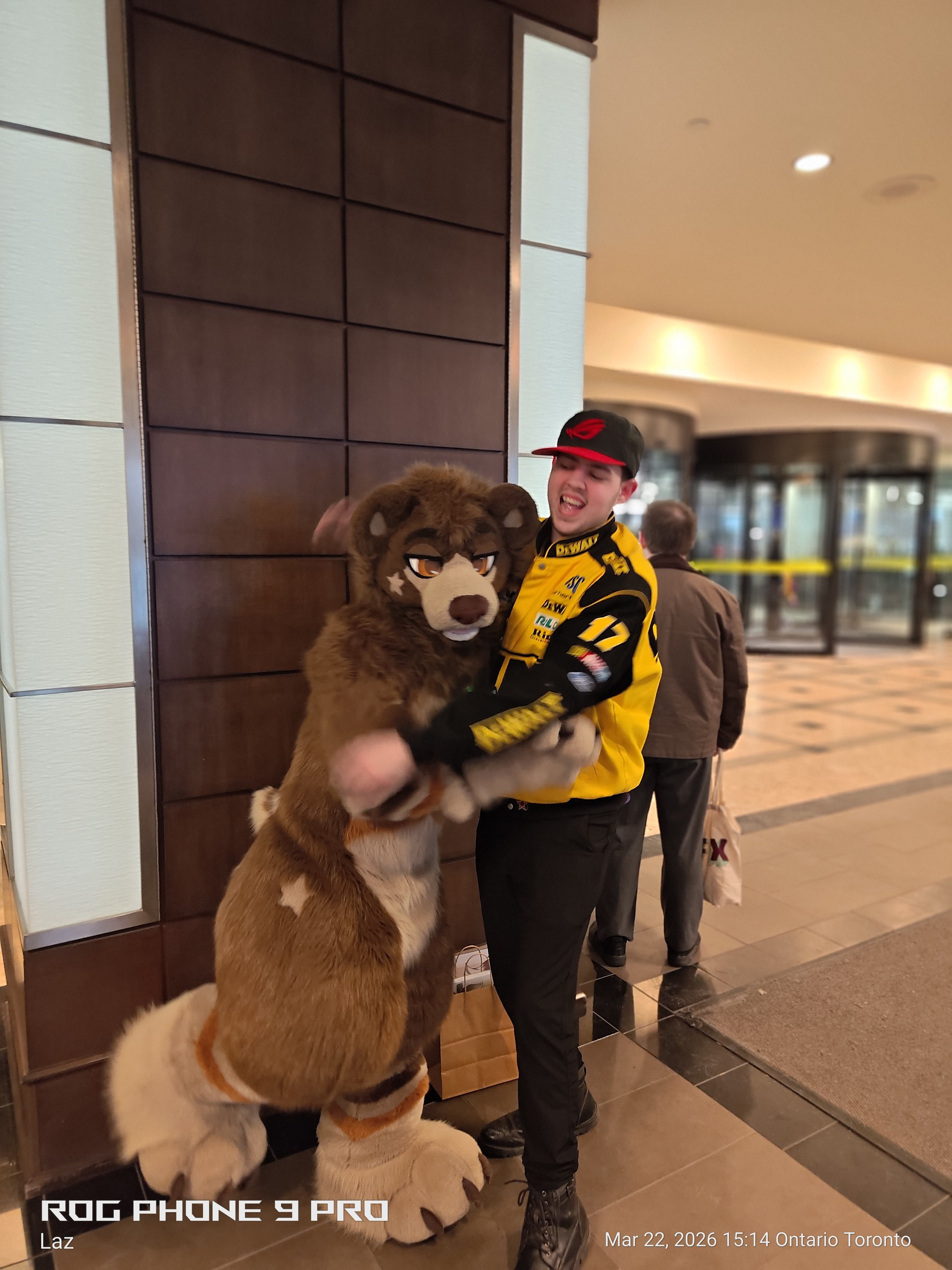A man in a yellow and black racing jacket is hugging a person in a lion costume, inside a building near an elevator.