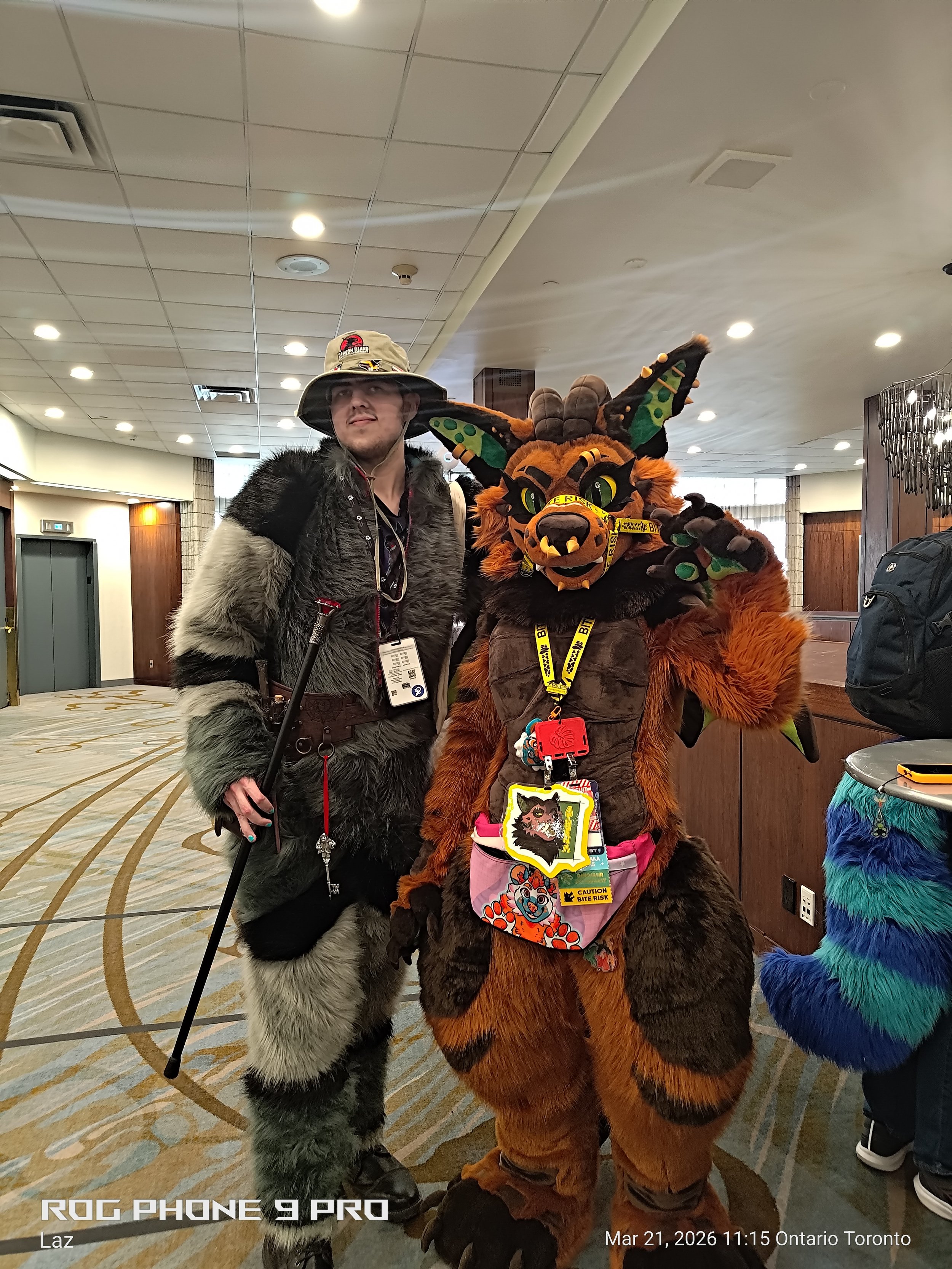 Two individuals in furry costumes posing together indoors at a convention. The person on the left is dressed in a gray and black wolf costume with a fur coat, hat, and walking stick. The person on the right is dressed in a vibrant orange and black mo