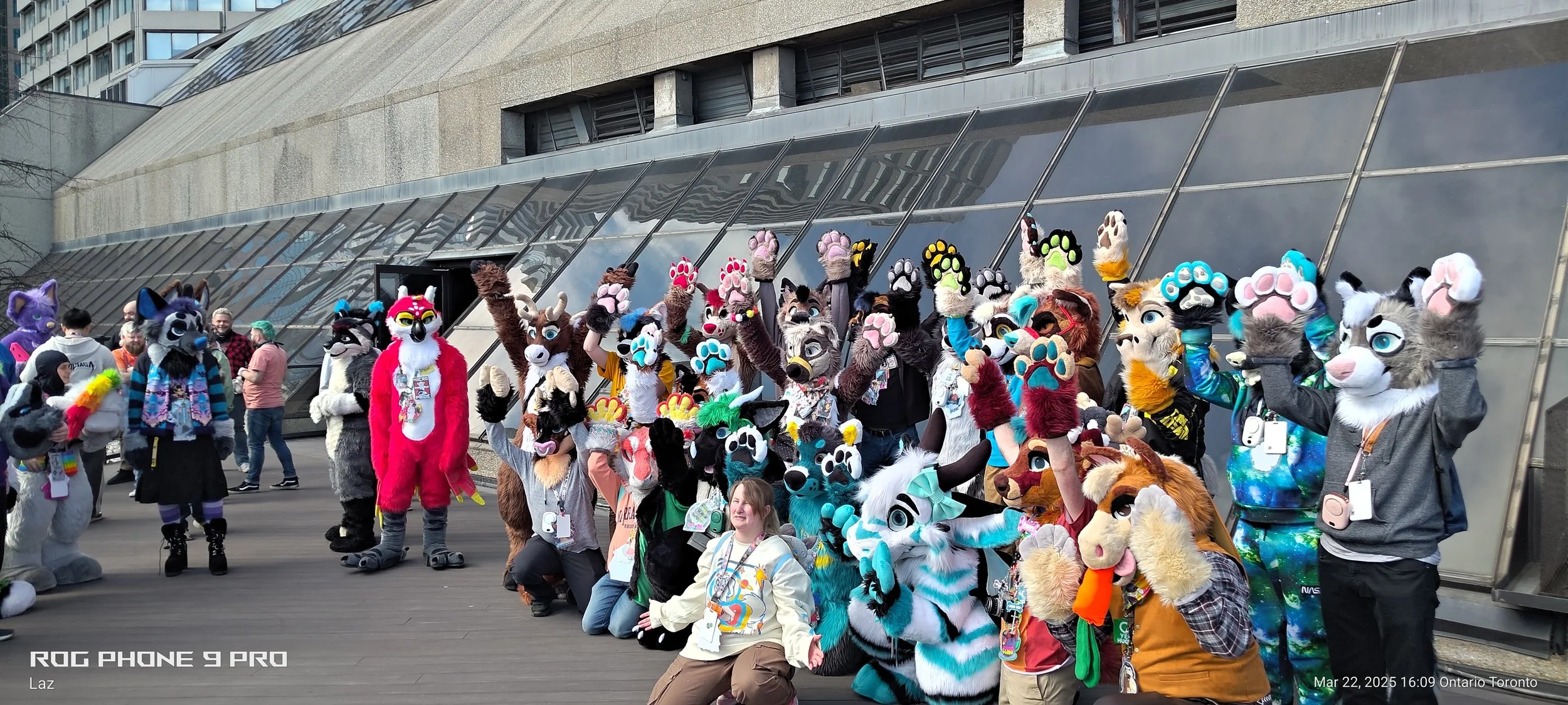 Group of people wearing colorful animal costumes and fursuits posing outdoors in front of a modern building with glass and concrete architecture.