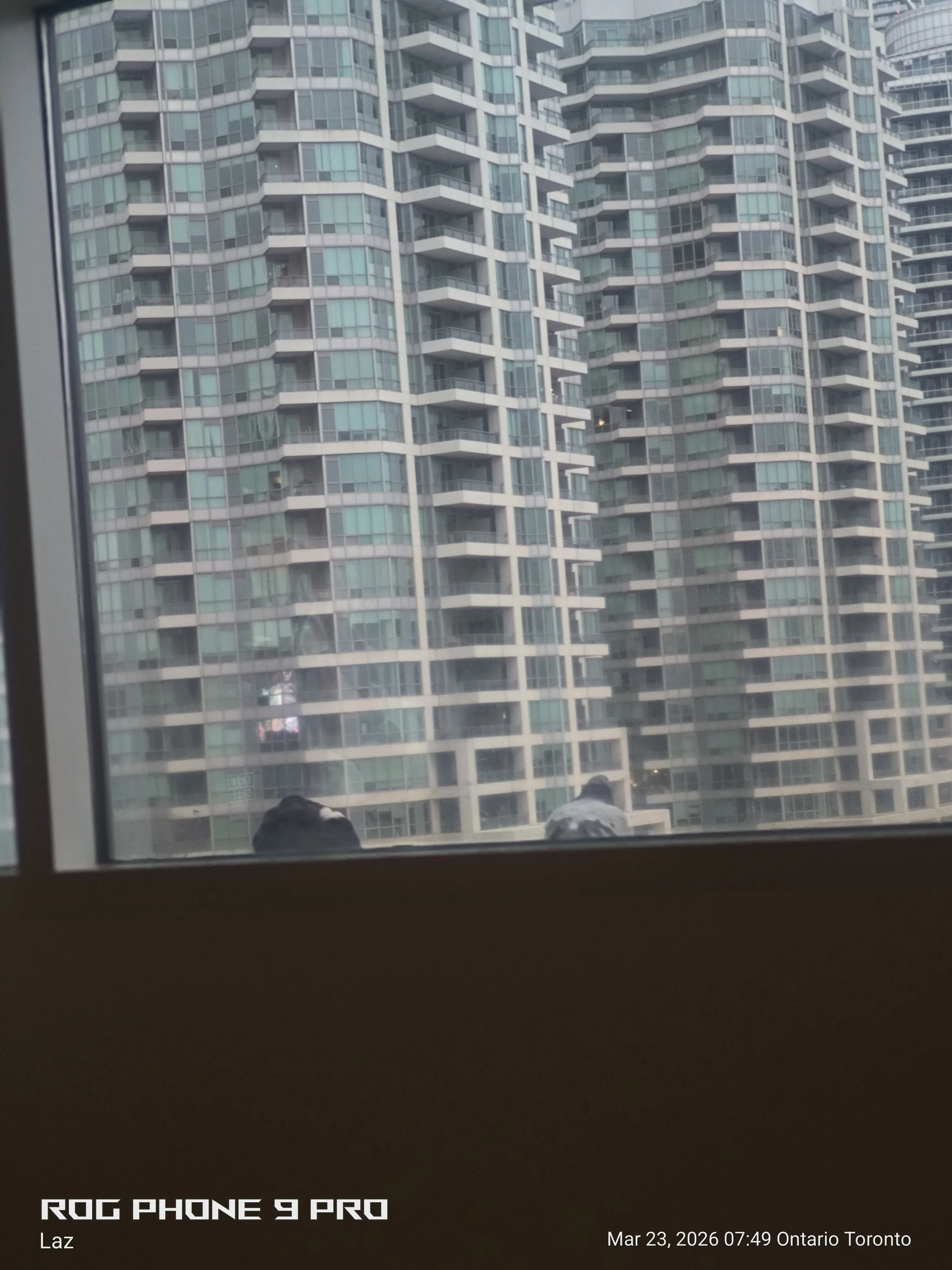 View of tall modern apartment buildings with glass balconies through a window. The photo appears to be taken from inside a room, with part of the window frame and a street view reflection visible. The text on the image indicates it was taken with a R