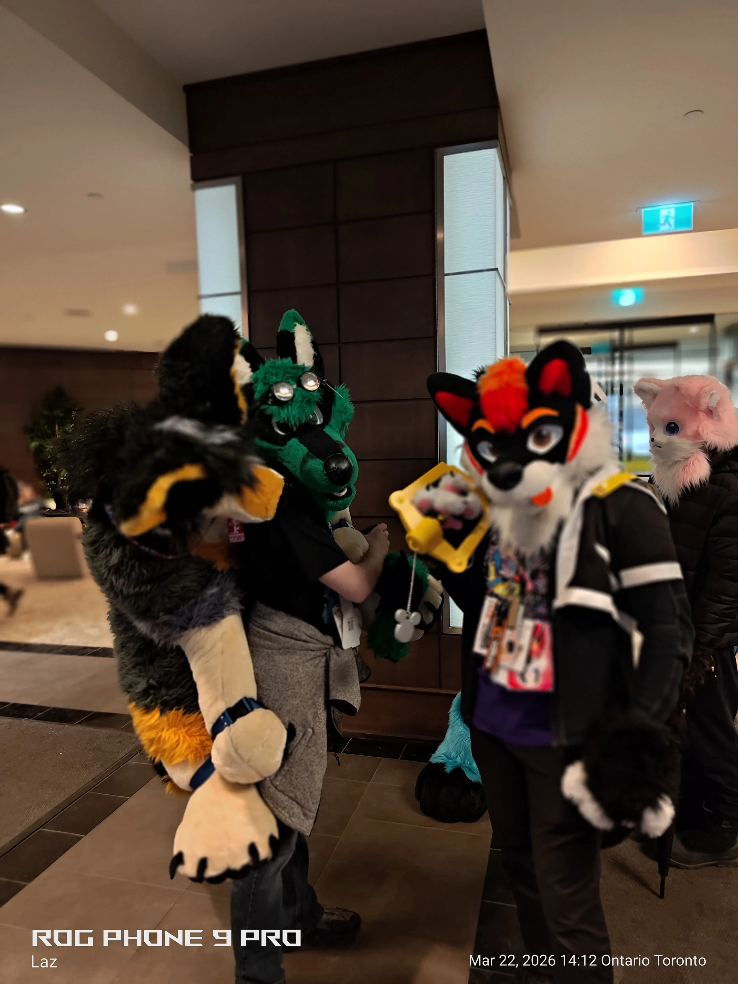 People in animal costumes socializing indoors, with one person in a green and black wolf costume and others dressed as a fox and a cat, in a large, well-lit space, possibly a hotel lobby or conference center.