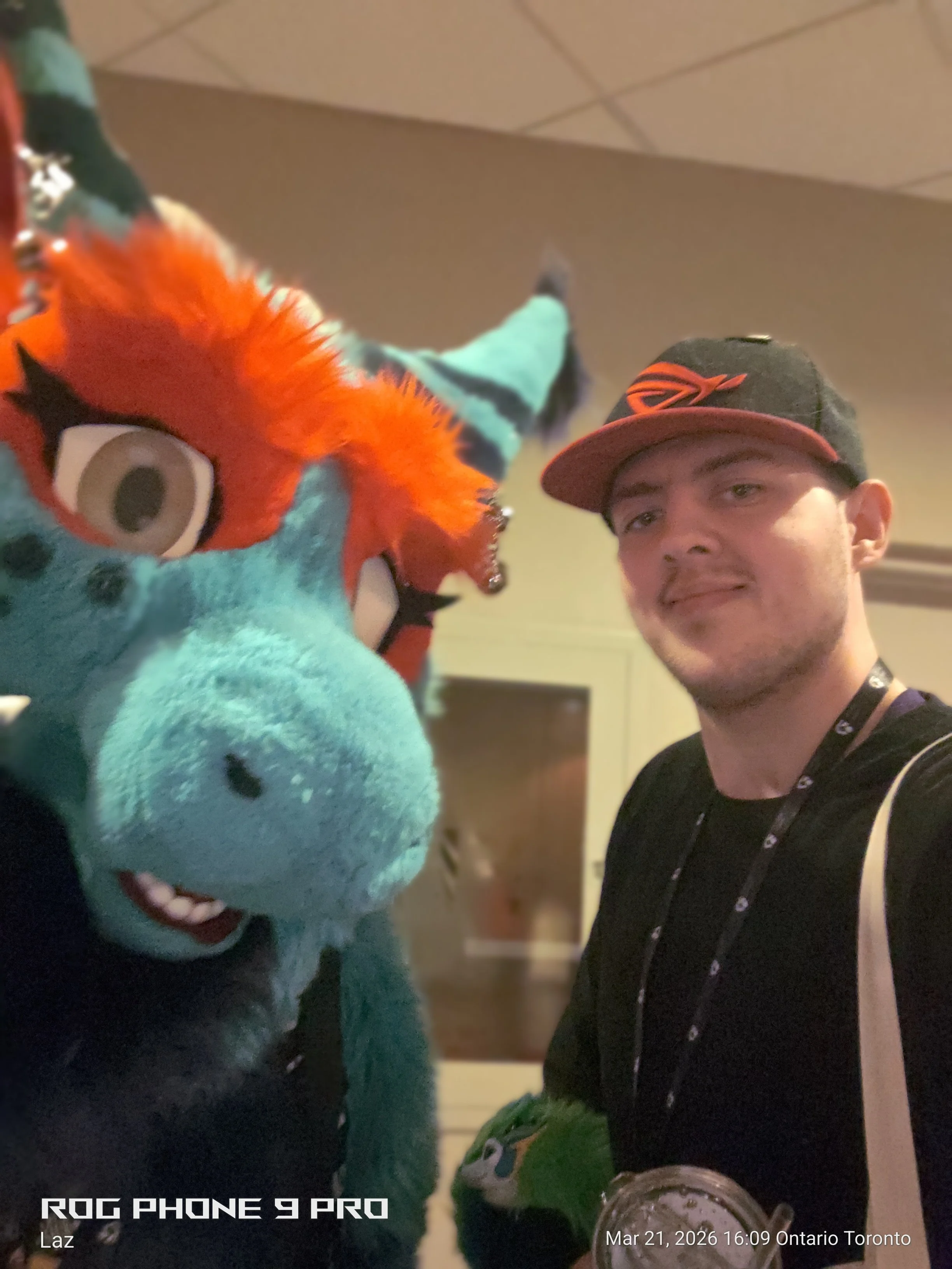 A person taking a selfie with a colorful furry creature costume resembling a dragon or dinosaur, and another person holding a drink in an indoor setting.